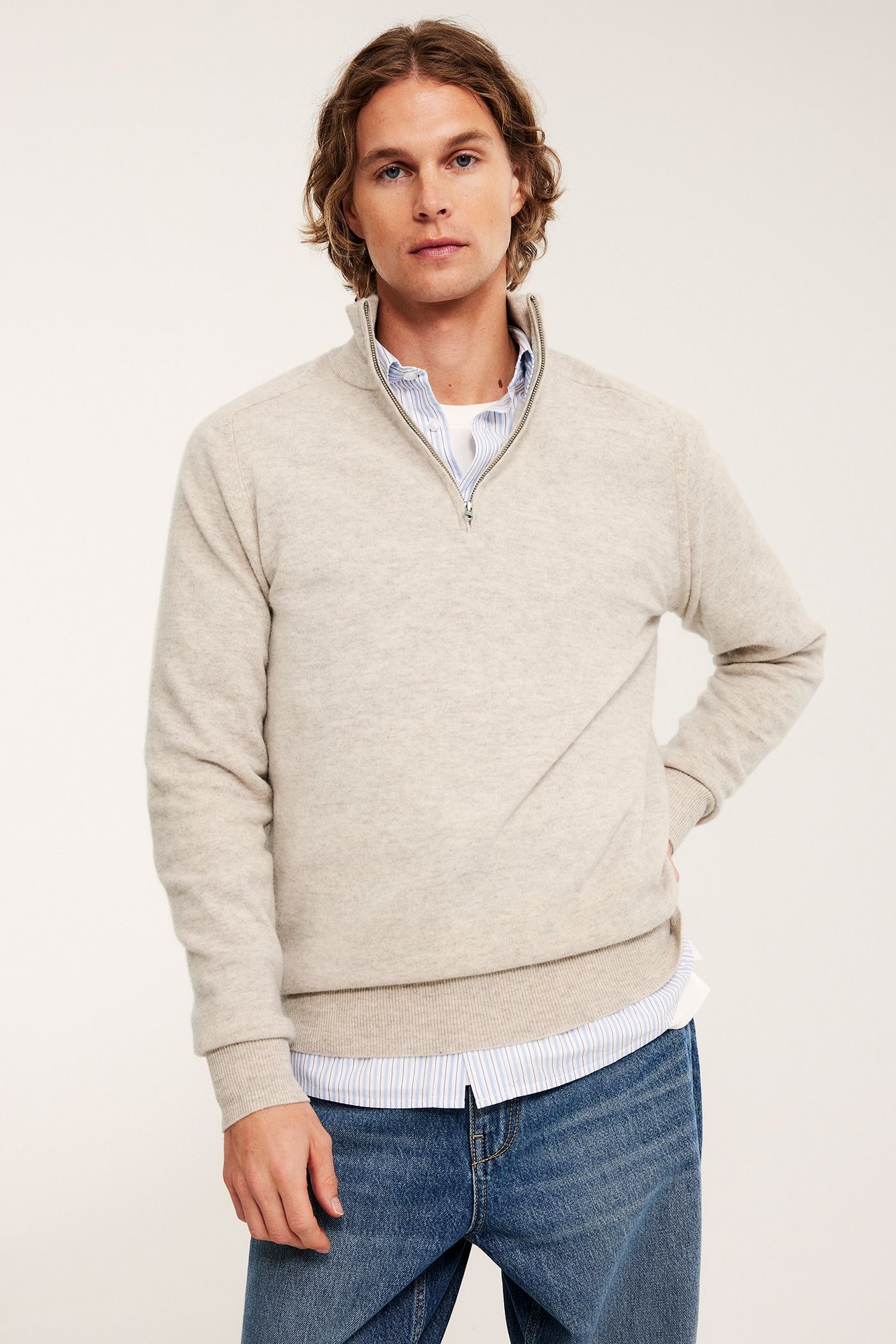 Frontbilde av mann i en beigemelert half-zip ullgenser over en stripete skjorte og blå jeans.