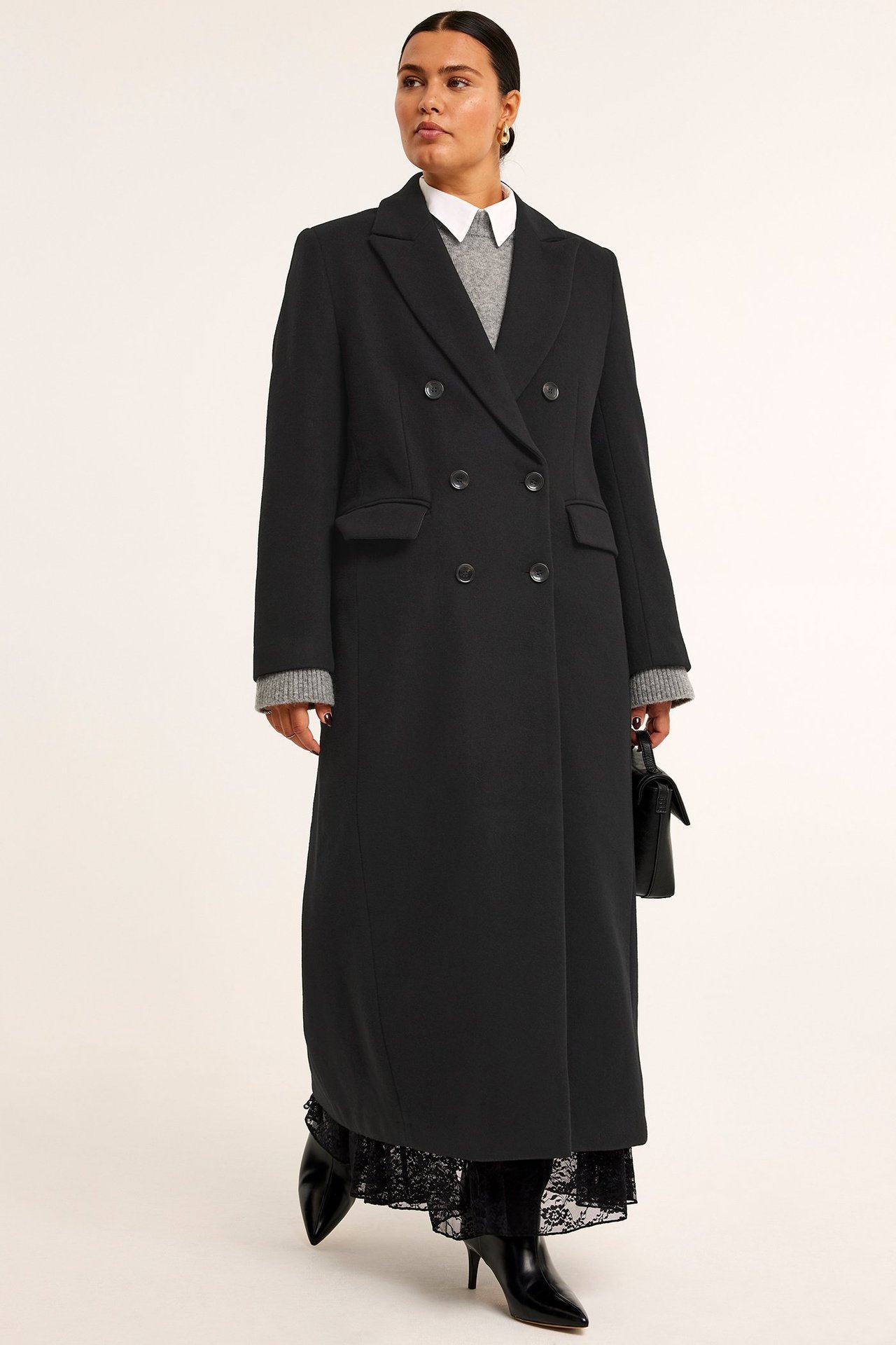 Nainen mustassa pitkässä takissa, harmaassa neuleessa, valkoisessa paidassa, mustassa hameessa, saappaissa, musta laukku.