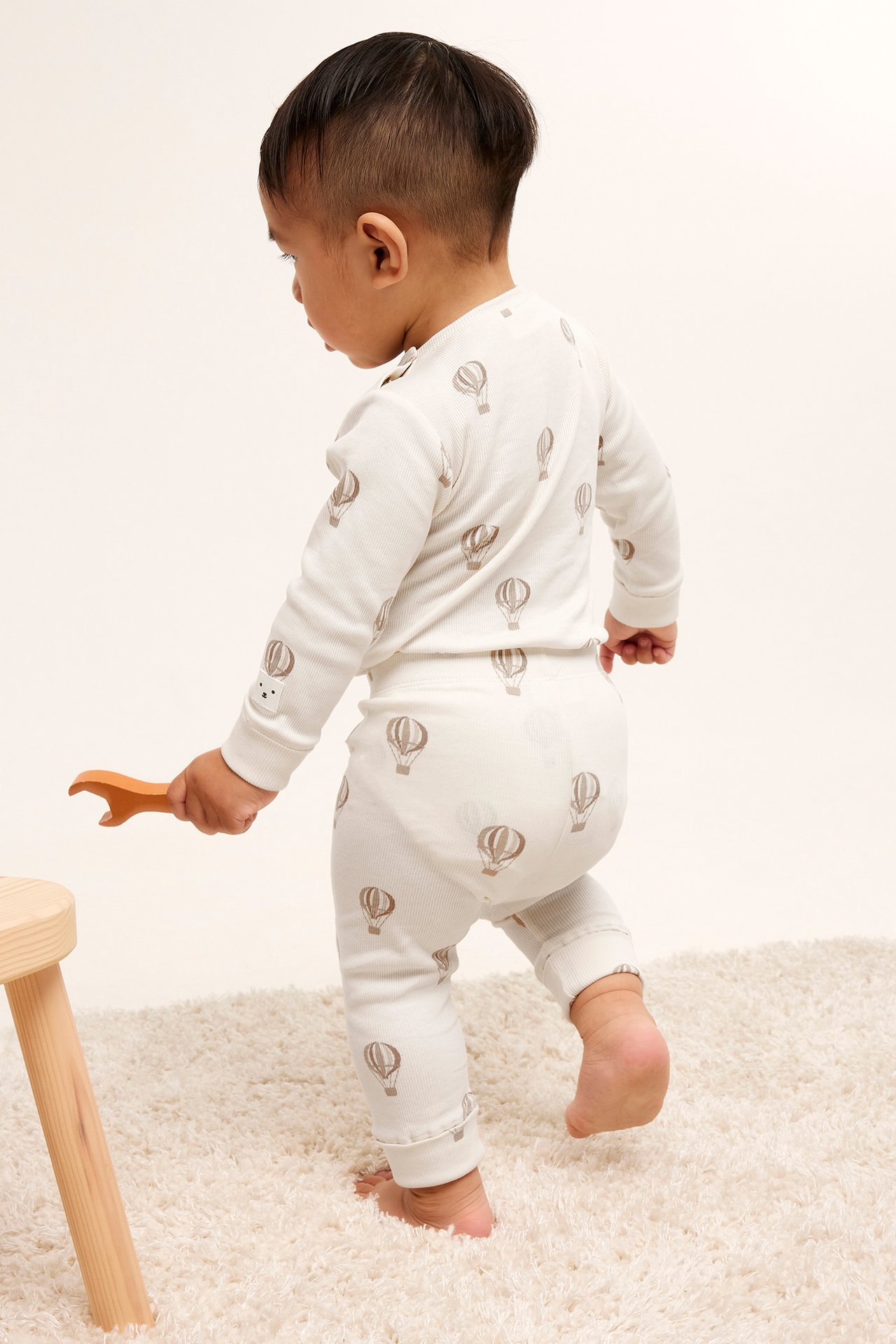 Bakifrån, baby i beige ribbat set med luftballongsmönster, håller en träleksak.