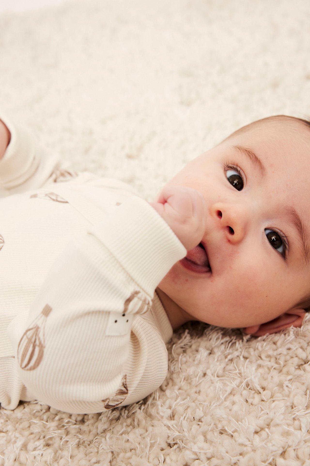 Lähikuva vauvasta beigessä ribbipaidassa, jossa ilmapallokuvio, käsi suun lähellä.