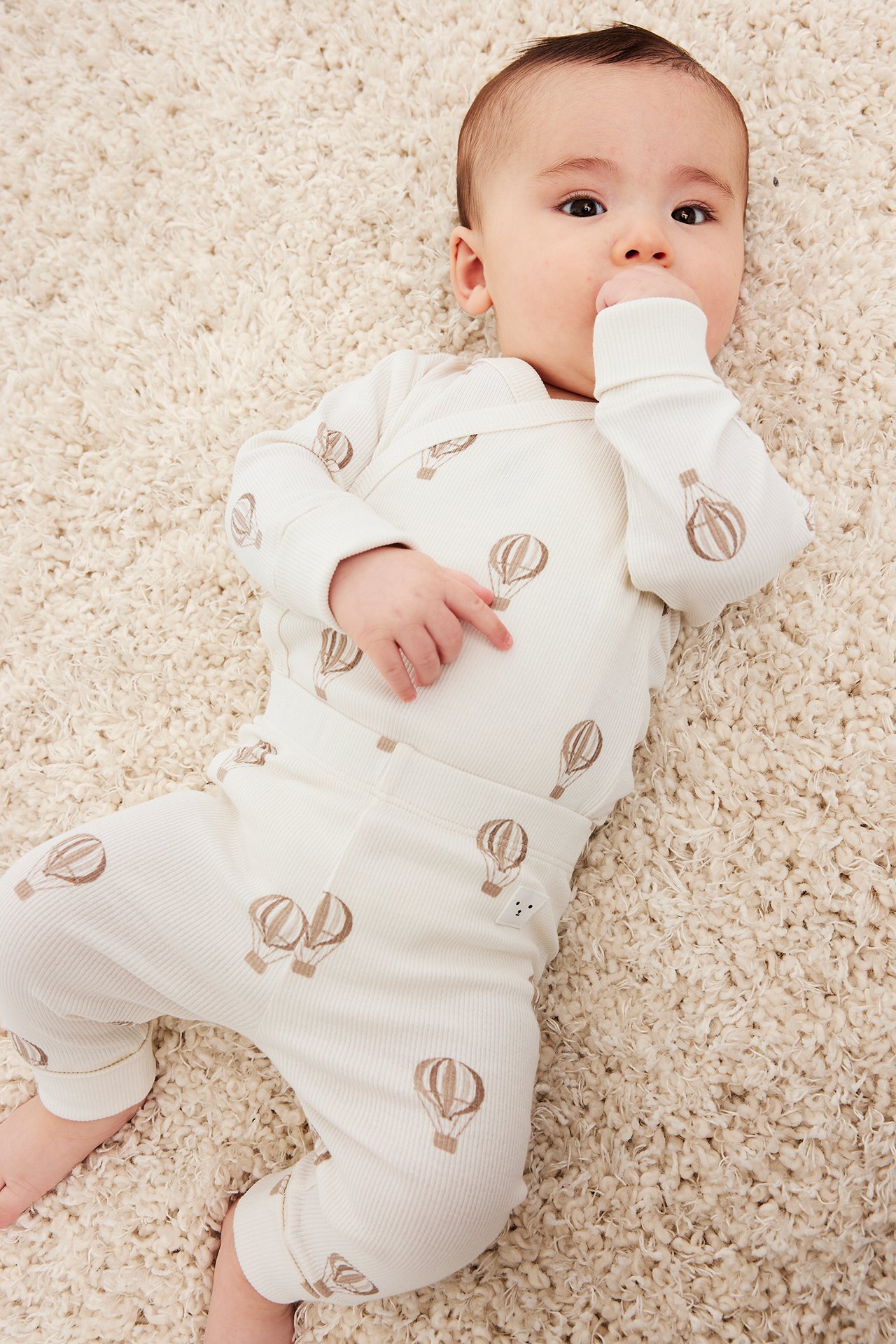 Ylhäältä näkymä: vauva beigessä ribbipotkupuvussa ja yhteensopivissa leggingseissä ruskeilla ilmapalloilla.