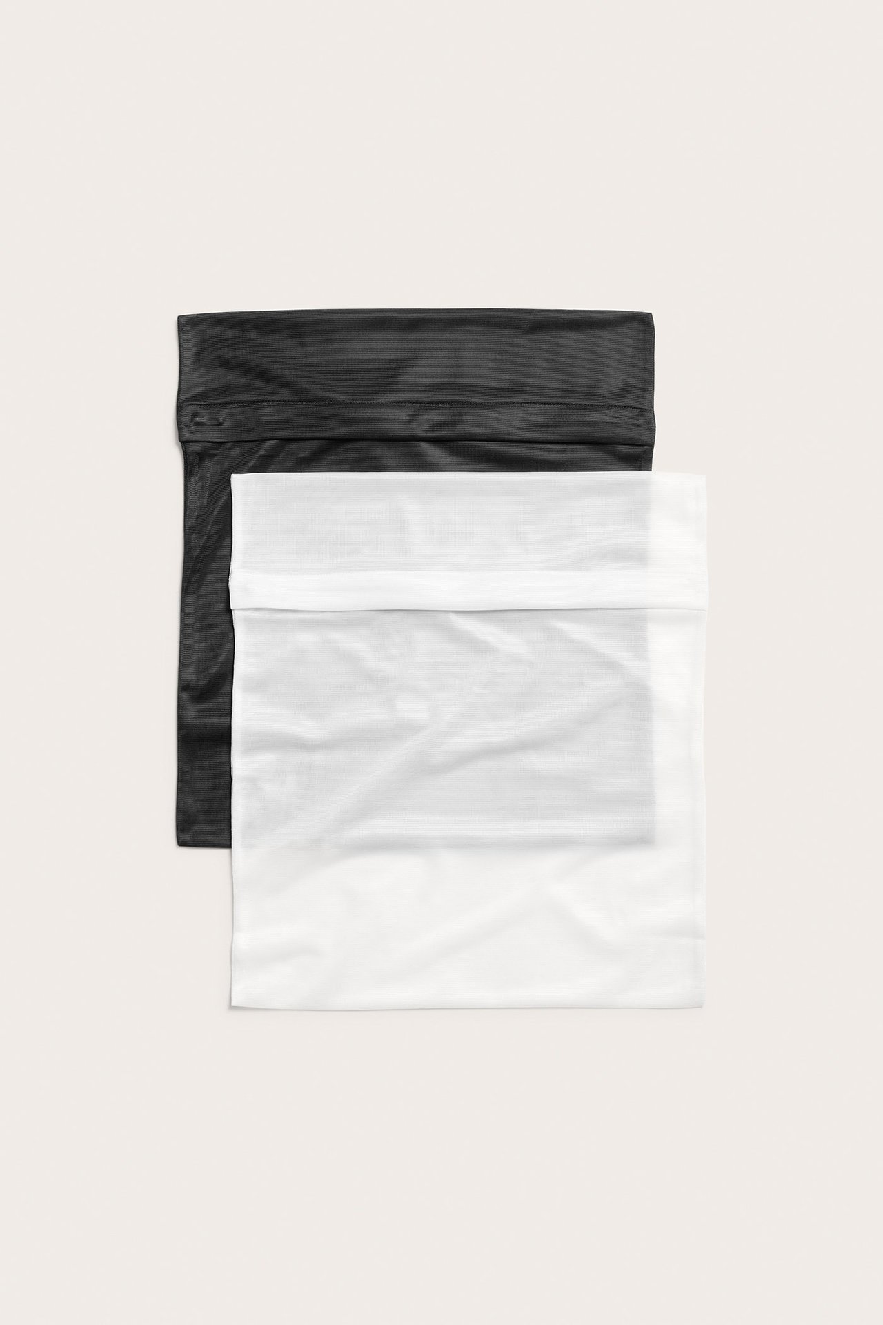 Framifrån, en vit tvättpåse i polyester med dold dragkedja, staplad över en svart tvättpåse.