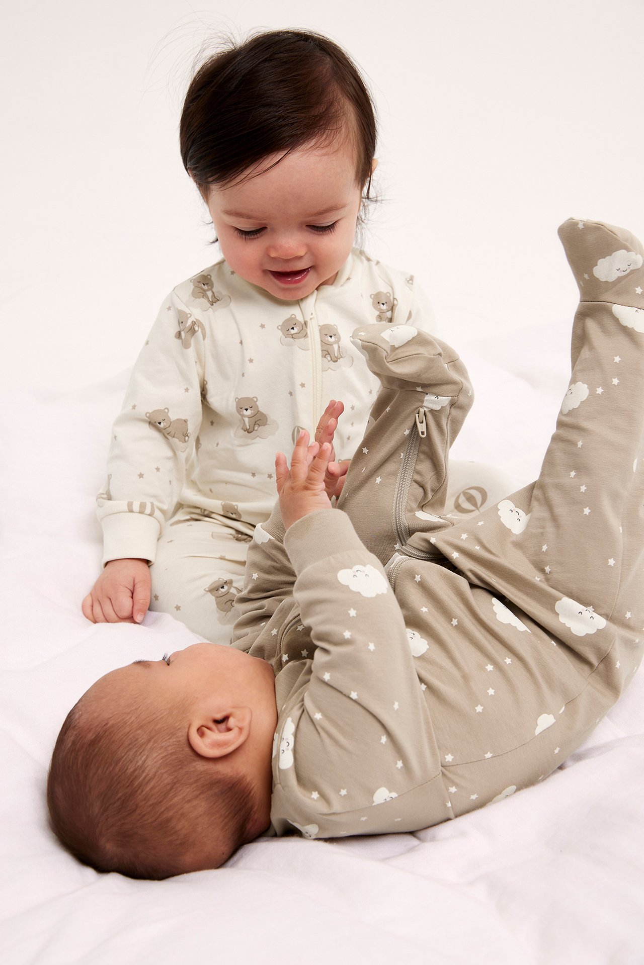 Framifrån: baby i benvit nallemönstrad pyjamas med dragkedja, en annan i ljusbrun molnmönstrad pyjamas.