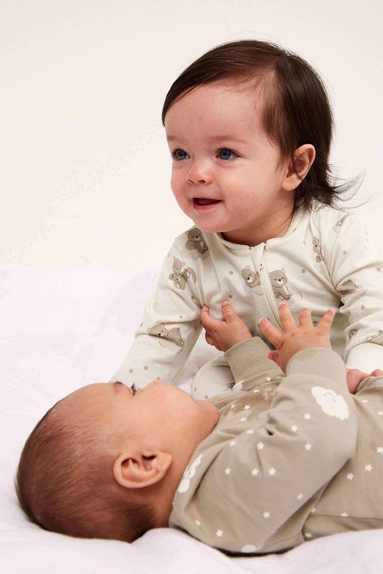 Framifrån: baby i offwhite björnmönstrad pyjamas med dragkedja, bredvid baby i beige stjärnpyjamas.