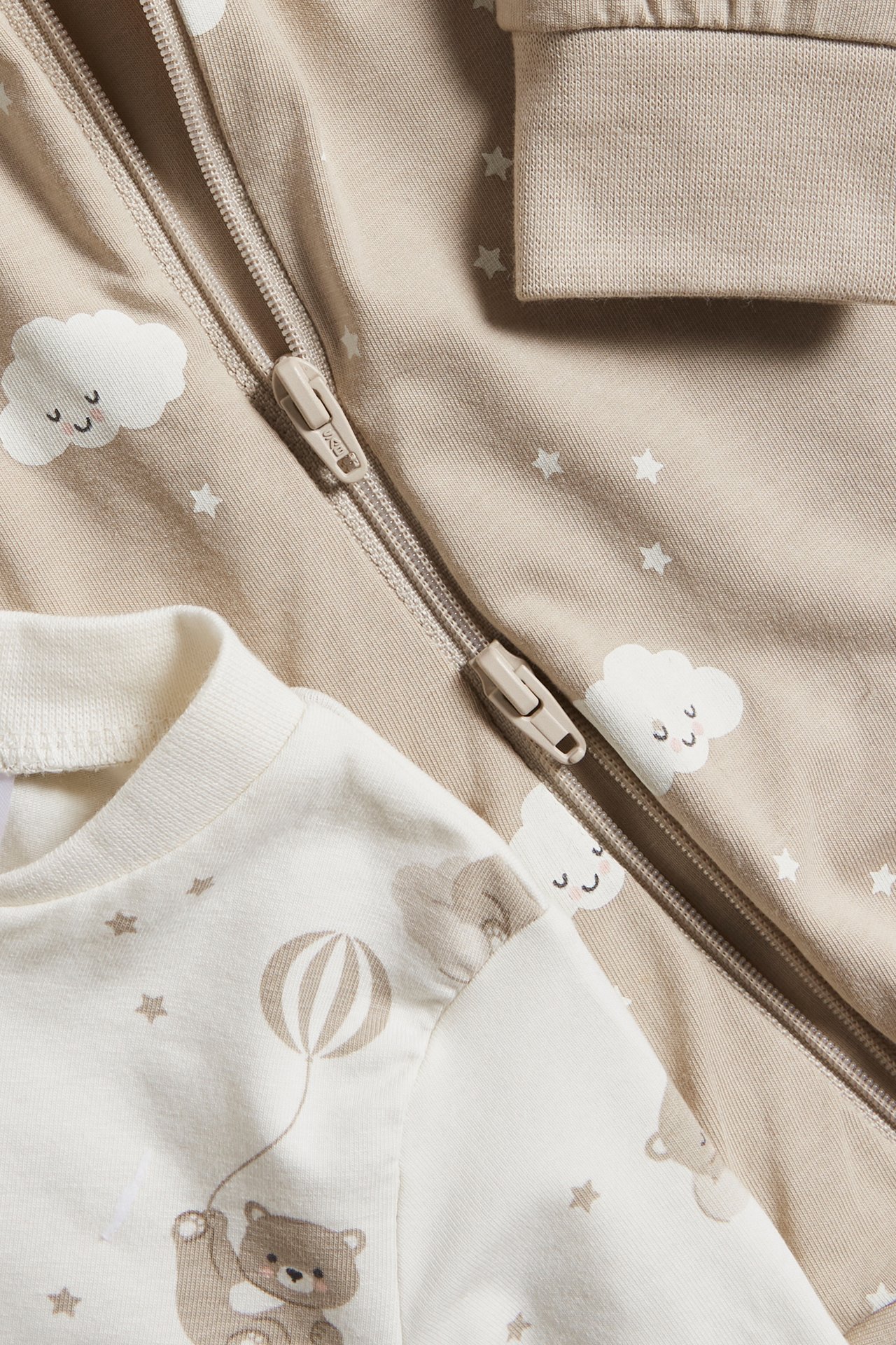 Närbild: två baby pyjamasar. Beige med moln/stjärnmönster, tvåvägsdragkedja. Off-white med nallebjörnsmönster.