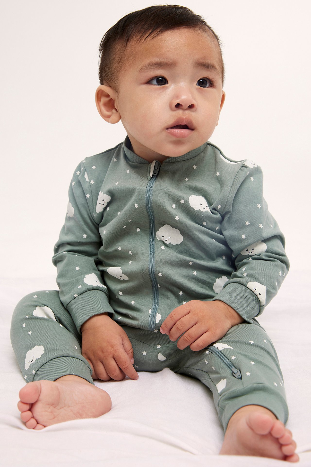 Framifrån, baby i ljusgrön mönstrad pyjamas med vita moln, stjärnor och tvåvägs dragkedja.