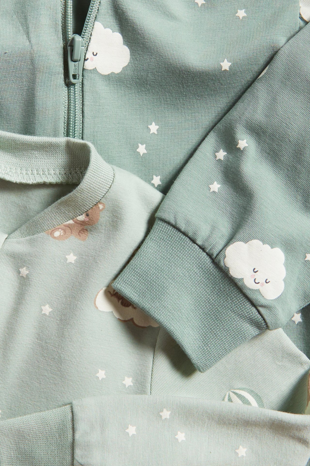 Närbild av två ljusgröna bomulls-pyjamasar för baby med vita moln- och stjärnmönster, en med dragkedja.