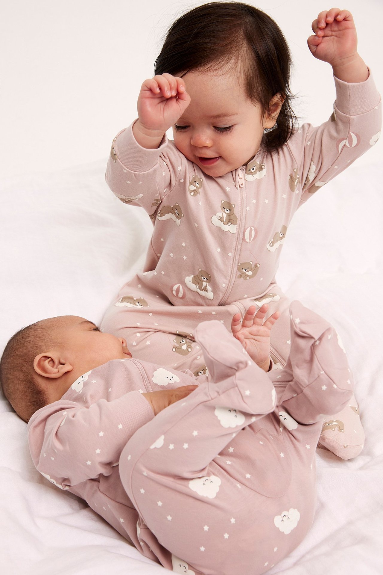 Edestä: kaksi vauvaa vaaleanpunaisissa kuviollisissa puuvillahaalareissa, joissa on jalkaosat ja kaksisuuntainen vetoketju.