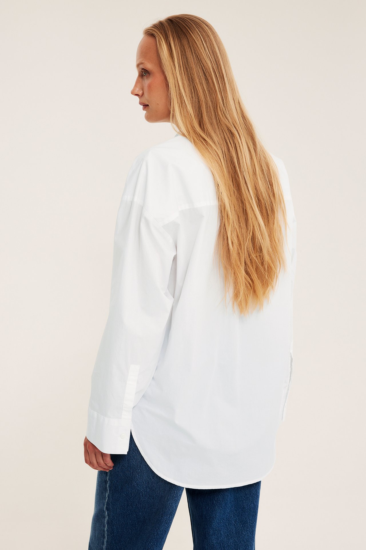 Sidebilde av en kvinne i en hvit oversized bomullspoplin-skjorte med avrundet kant og blå jeans.