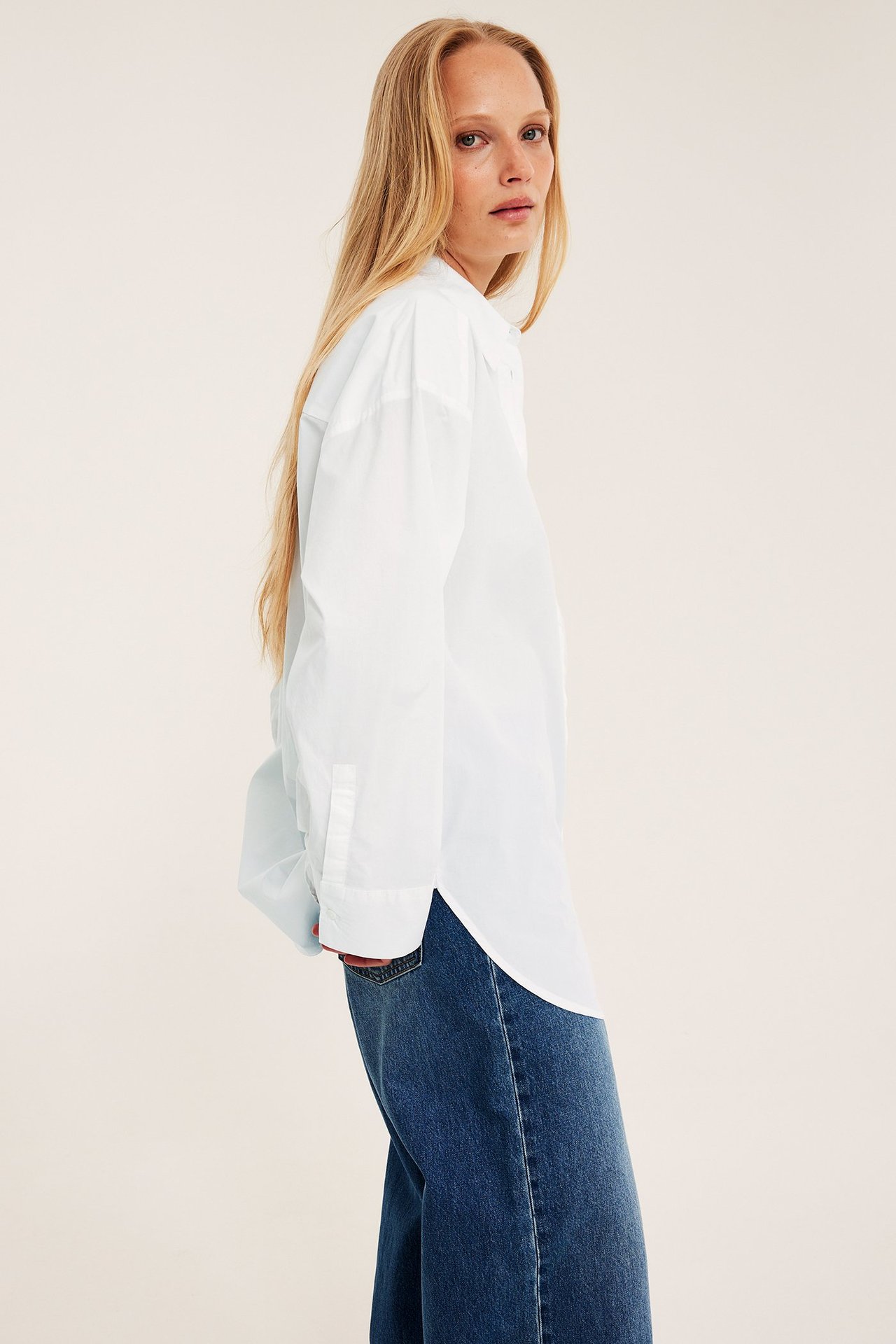 Sidebilde av en kvinne i en hvit oversized bomullspoplin-skjorte og mørkeblå jeans.