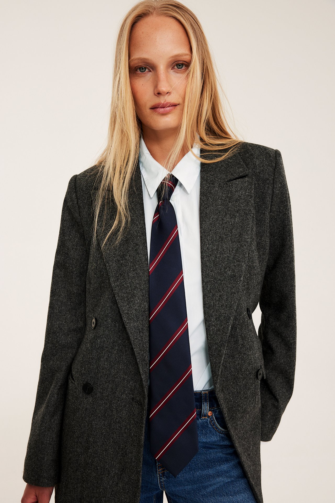 Frontvisning: Kvinne i lys blå oversized poplinskjorte, mørkegrå blazer, marineblått stripete slips og blå jeans.