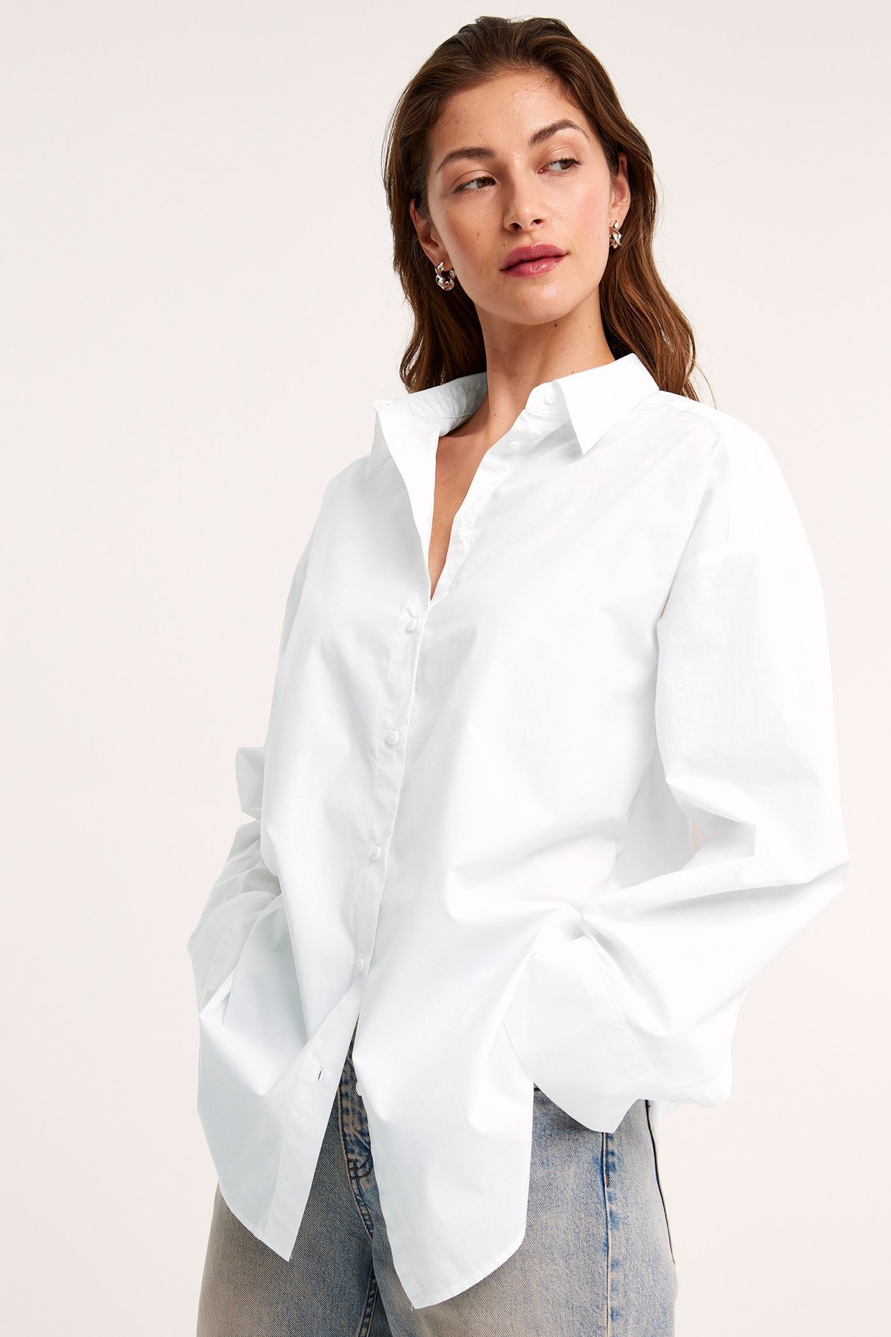 Edestä, nainen yllään ylisuuri valkoinen puuvillapopliinipaita, vaaleansiniset farkut ja hopeiset korvarenkaat.