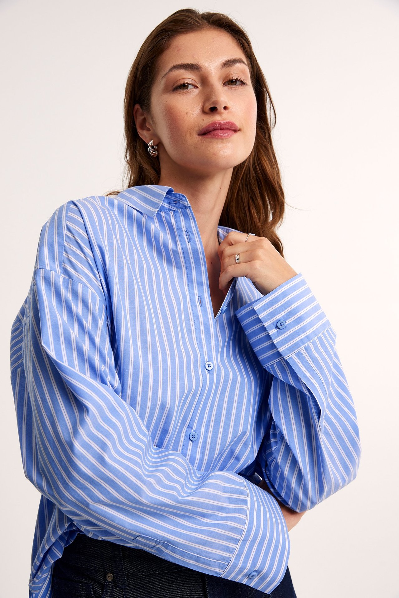 Nærbilde forfra av kvinne i oversized blå- og hvitstripet bomullspoplinskjorte, med sølvøreringer.