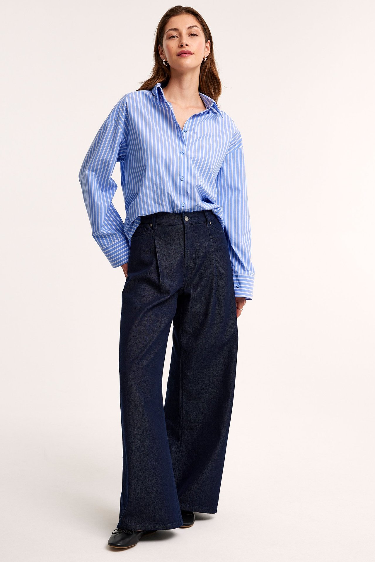 Forfra, kvinne i oversized blåstripet bomullsskjorte, mørkeblå vide bukser og svarte flate sko.