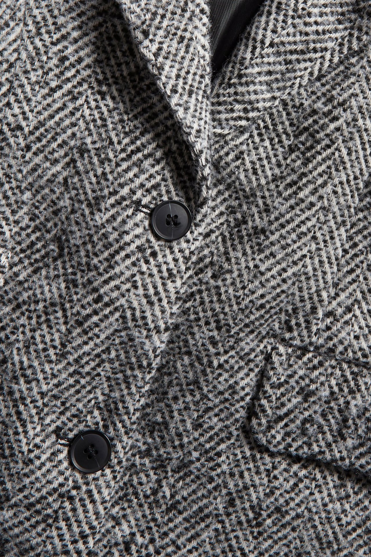 Zbliżenie czarno-białej marynarki tweedowej w jodełkę z dwoma czarnymi guzikami i kieszenią z klapką.