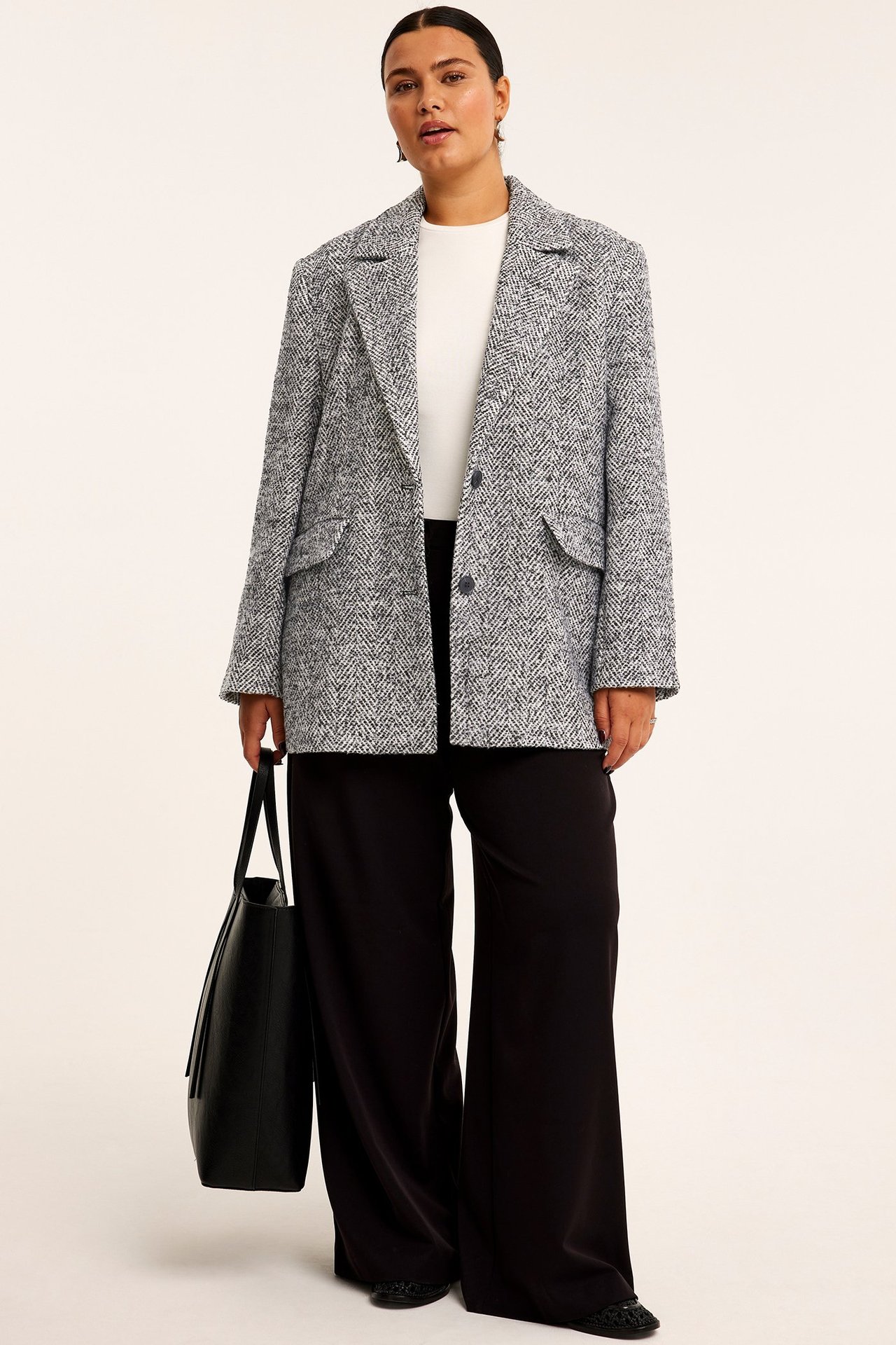 Widok z przodu: Kobieta w czarno-białej tweedowej marynarce, białym topie, czarnych spodniach, trzyma czarną torbę.
