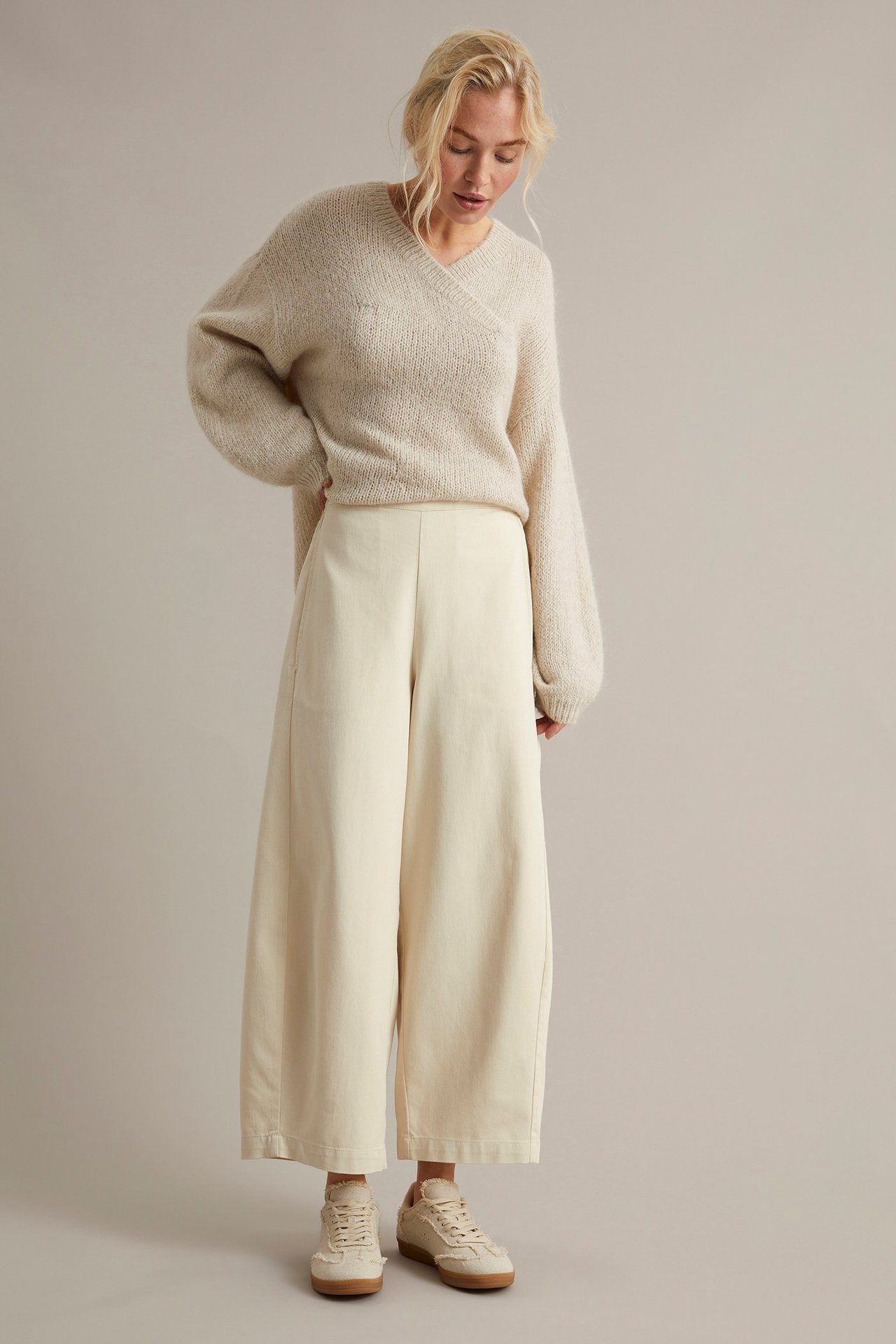 Edestäpäin: nainen yllään vaaleanbeiget leveälahkeiset pull on -housut, beige kietaisuneule ja lenkkarit.