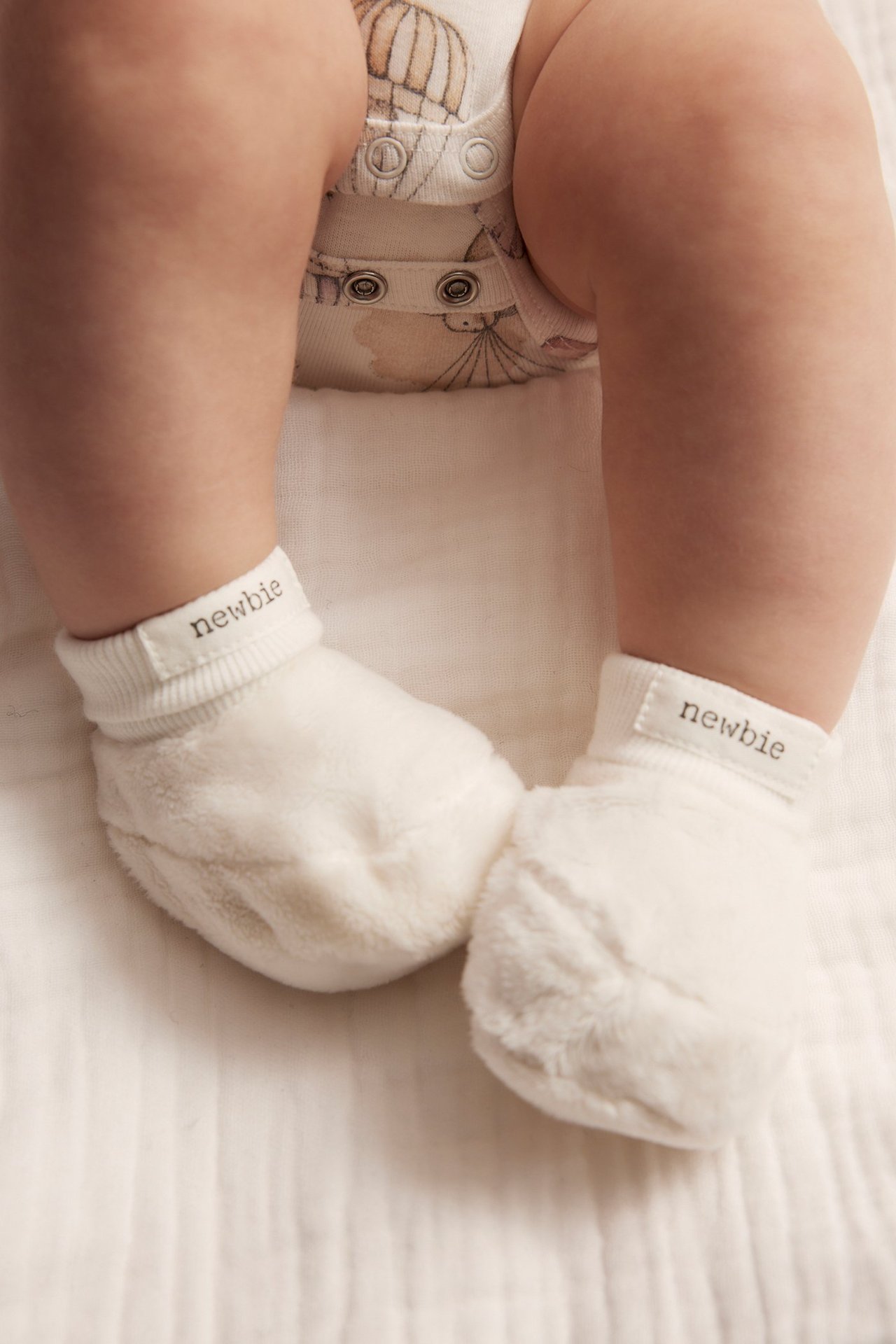 Närbild på babys fötter i offwhite fluffiga tossor med 'newbie' etiketter, över en mönstrad body.