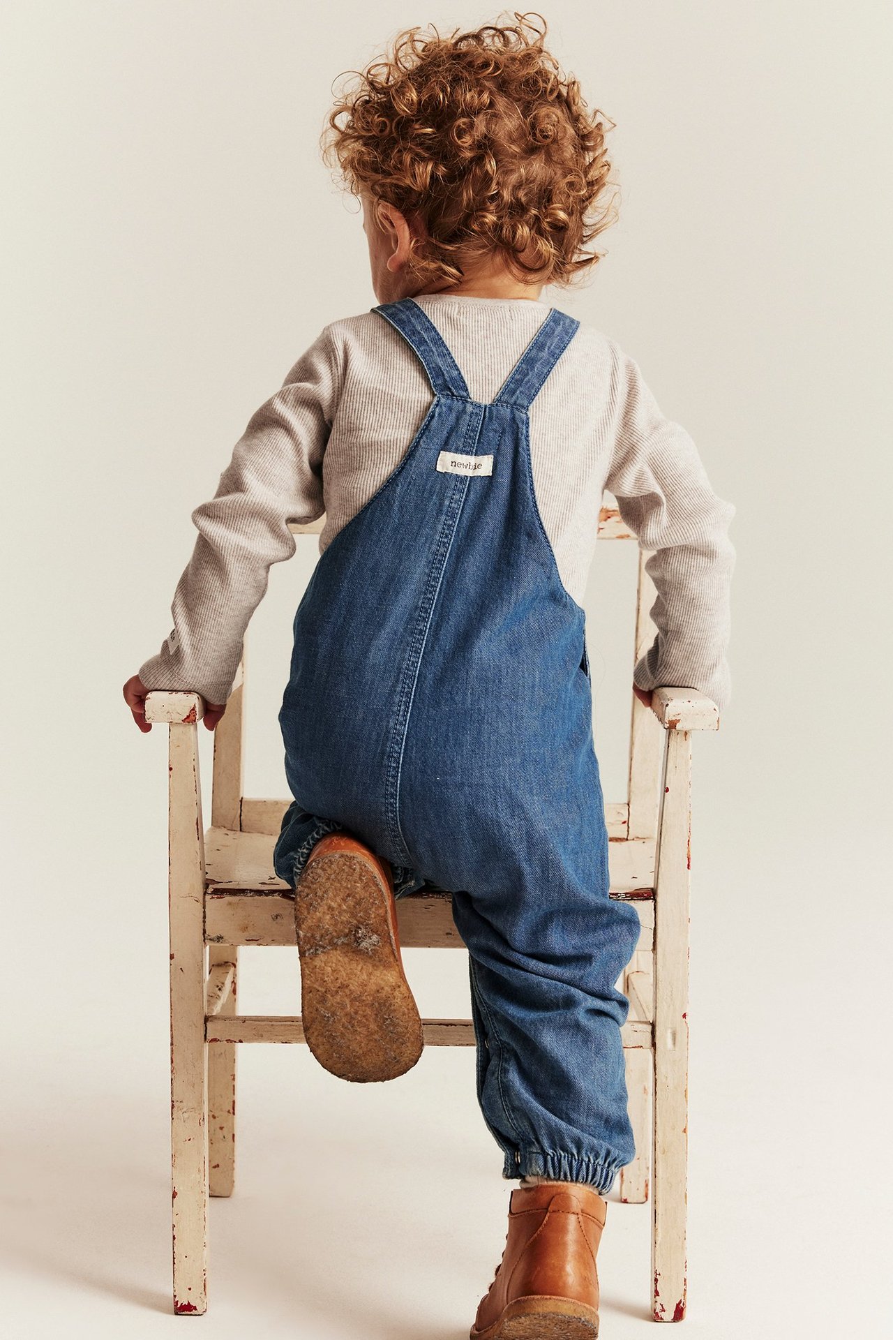 Ryggbilde av et barn i beige ribbet langermet body, blå denimoverall og brune støvler.