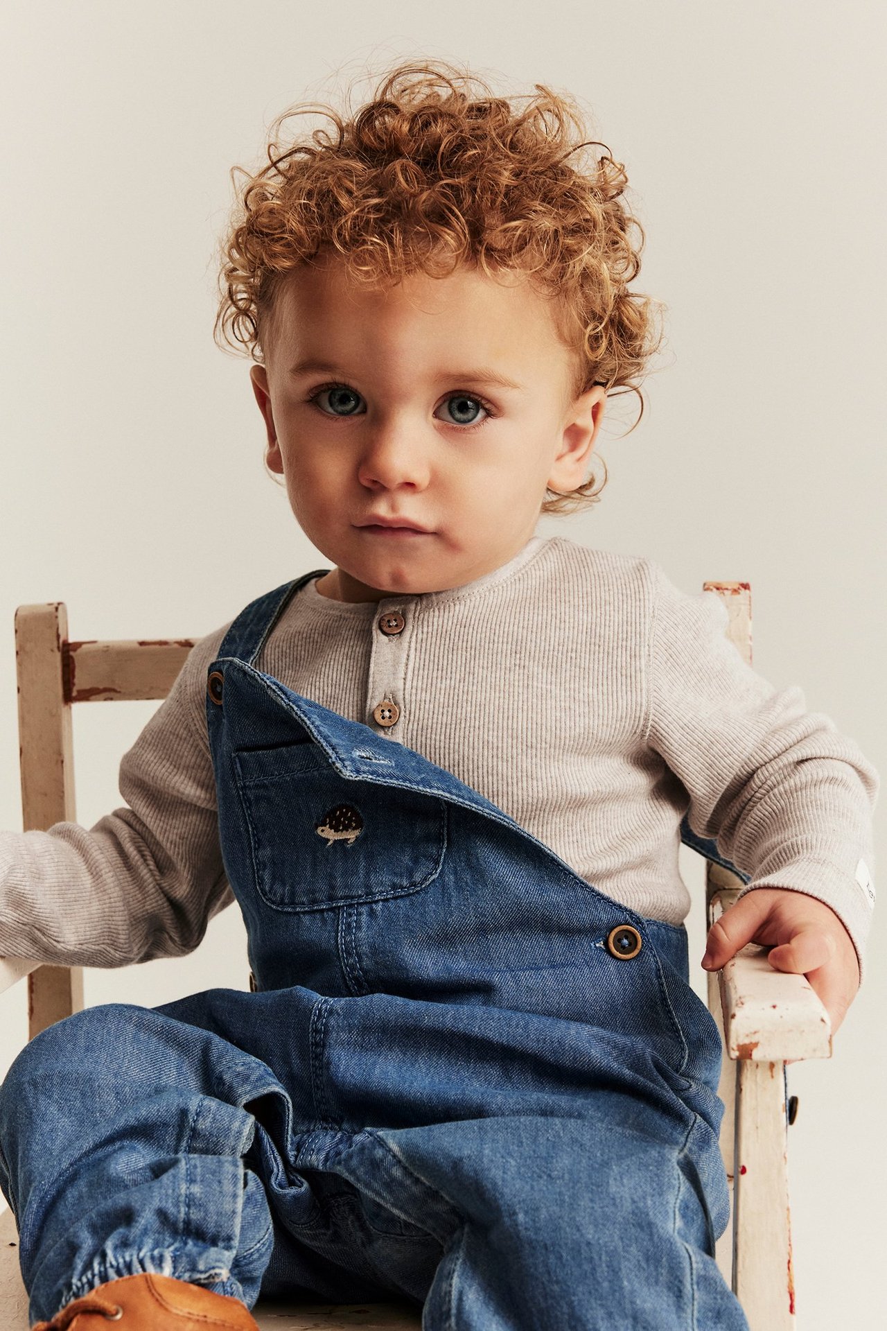 Frontutsikt av en baby i beige melange ribbet langermet body under blå denim-overall.