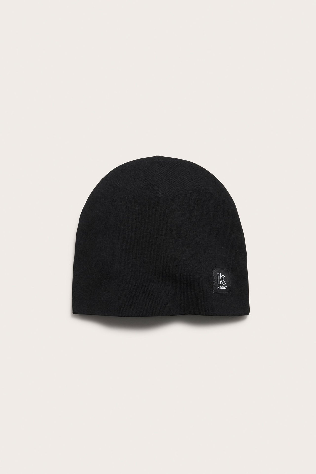 Framifrån, svart enfärgad trikåmössa med liten "k kaxs" etikett på sidan.