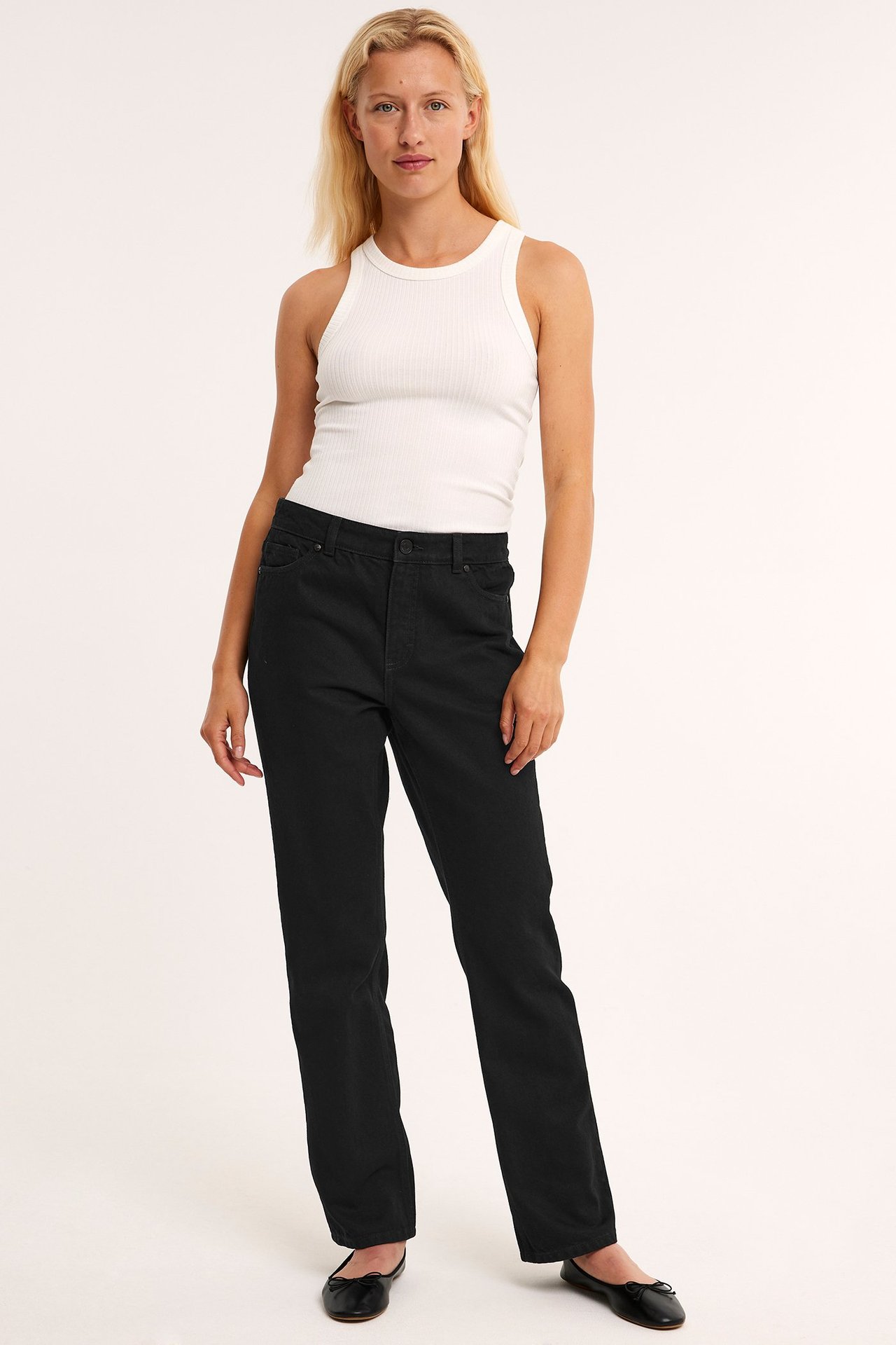 Frontvy av kvinna i svarta raka jeans med hög midja, vitt ribbat linne och svarta ballerinaskor.