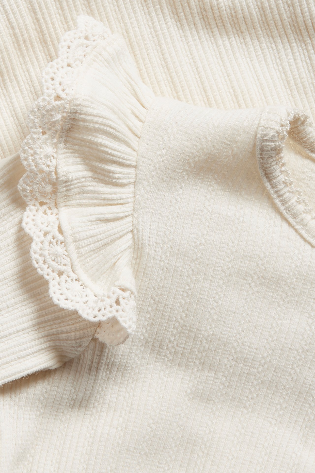 Nærbilde av en offwhite ribbet babybody med lange ermer, volangskuldre og blondekant.