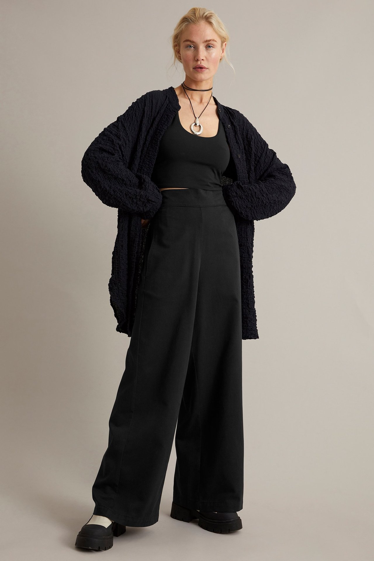 Forfra: Kvinne i svarte vide pull on-bukser, svart singlet, teksturert cardigan og platåsko.