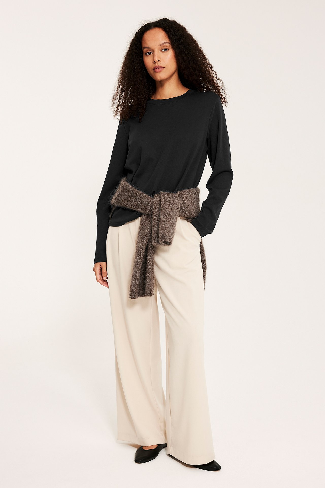 Widok z przodu: kobieta w czarnym bawełnianym topie z długim rękawem, kremowych spodniach, brązowy sweter zawiązany w pasie.