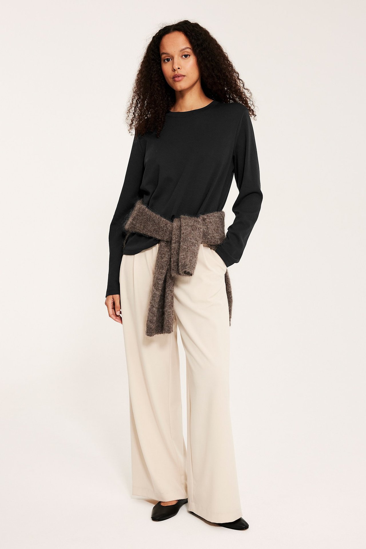 Forfra: kvinne i svart langermet bomullstopp, kremfargede vide bukser, brun genser knyttet rundt midjen.