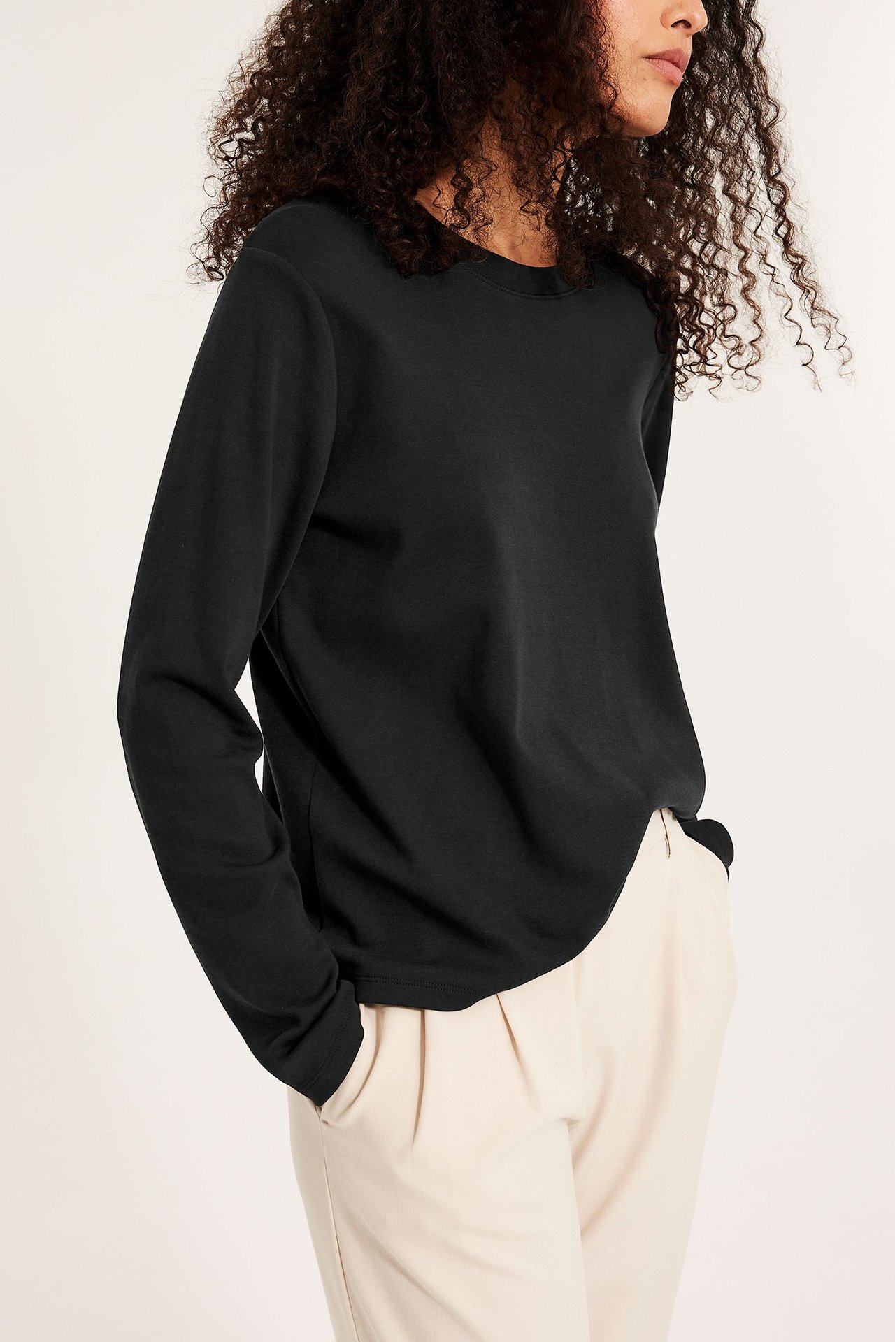 Frontvy av en kvinna i en svart långärmad bomullstopp och ljusbeige plisserade byxor.