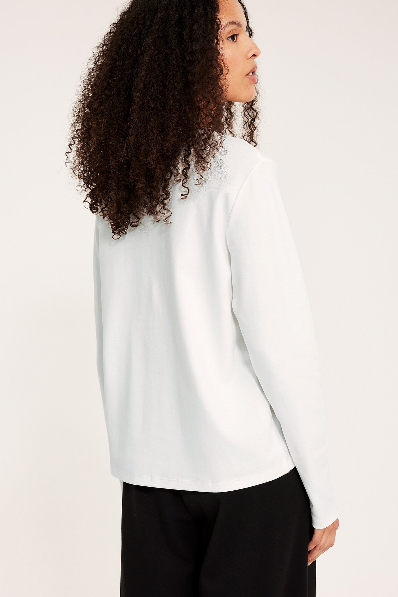 Naisen sivu-takakuva, yllään väljä luonnonvalkoinen pitkähihainen puuvillatoppi ja mustat housut.
