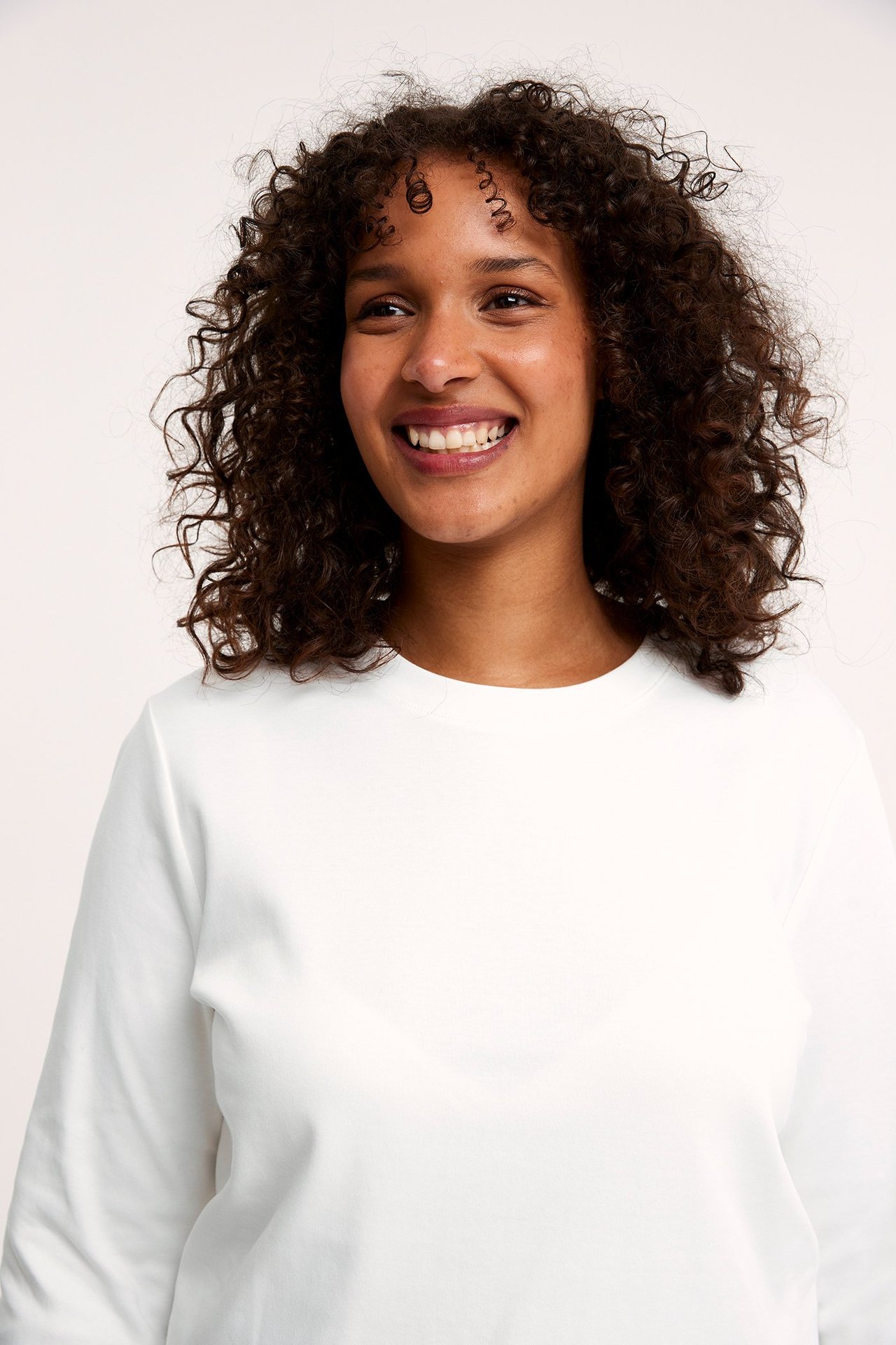 Nærbilde forfra av en smilende kvinne i en offwhite langermet t-skjorte med rund hals i bomull.