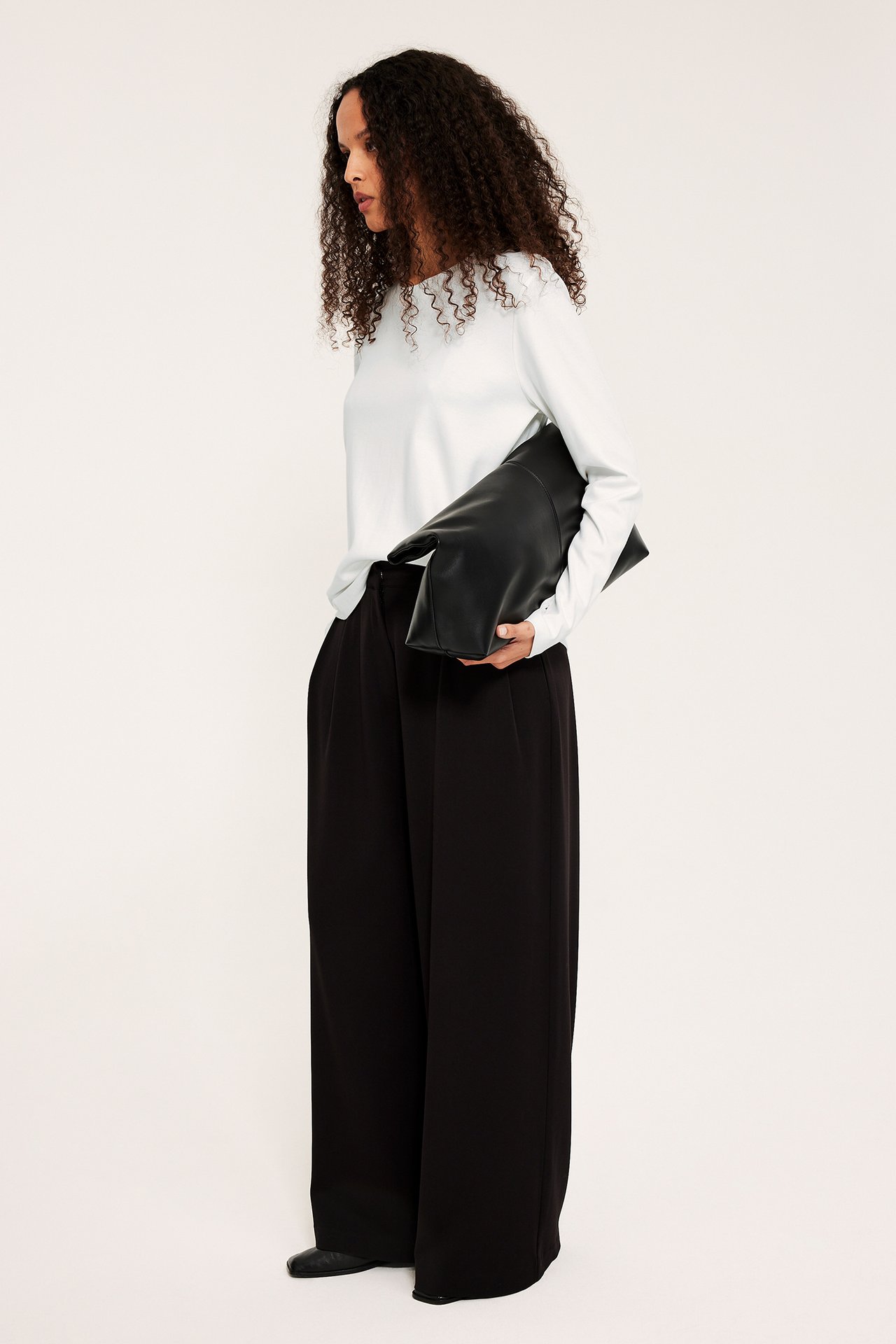 Sivukuva: Nainen yllään luonnonvalkoinen pitkähihainen puuvillatoppi, mustat leveät housut, pitää mustaa clutchia.