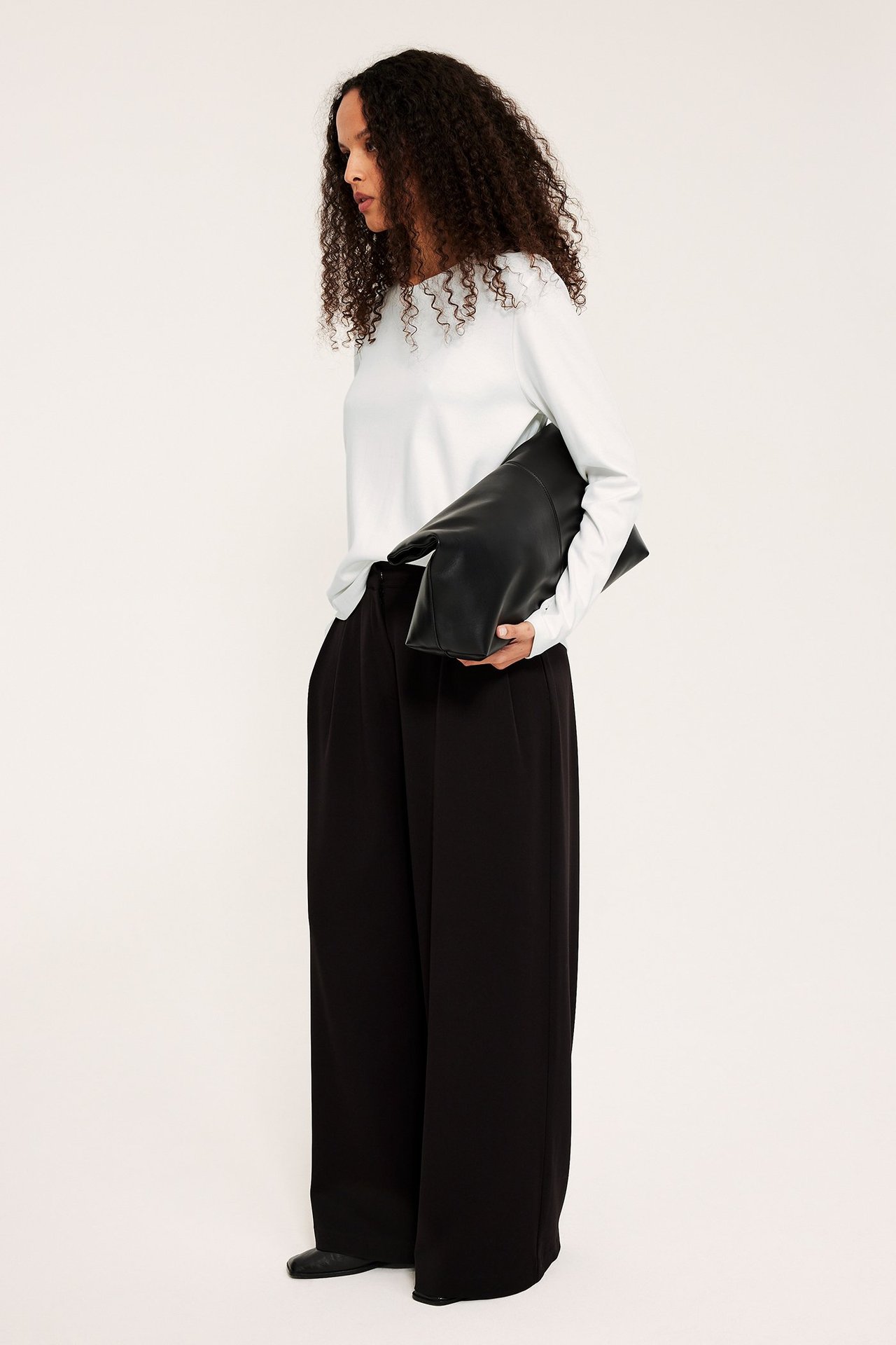Sideprofil: Kvinne bærer en offwhite langermet basistopp i bomull, svarte vide bukser, holder svart clutch.