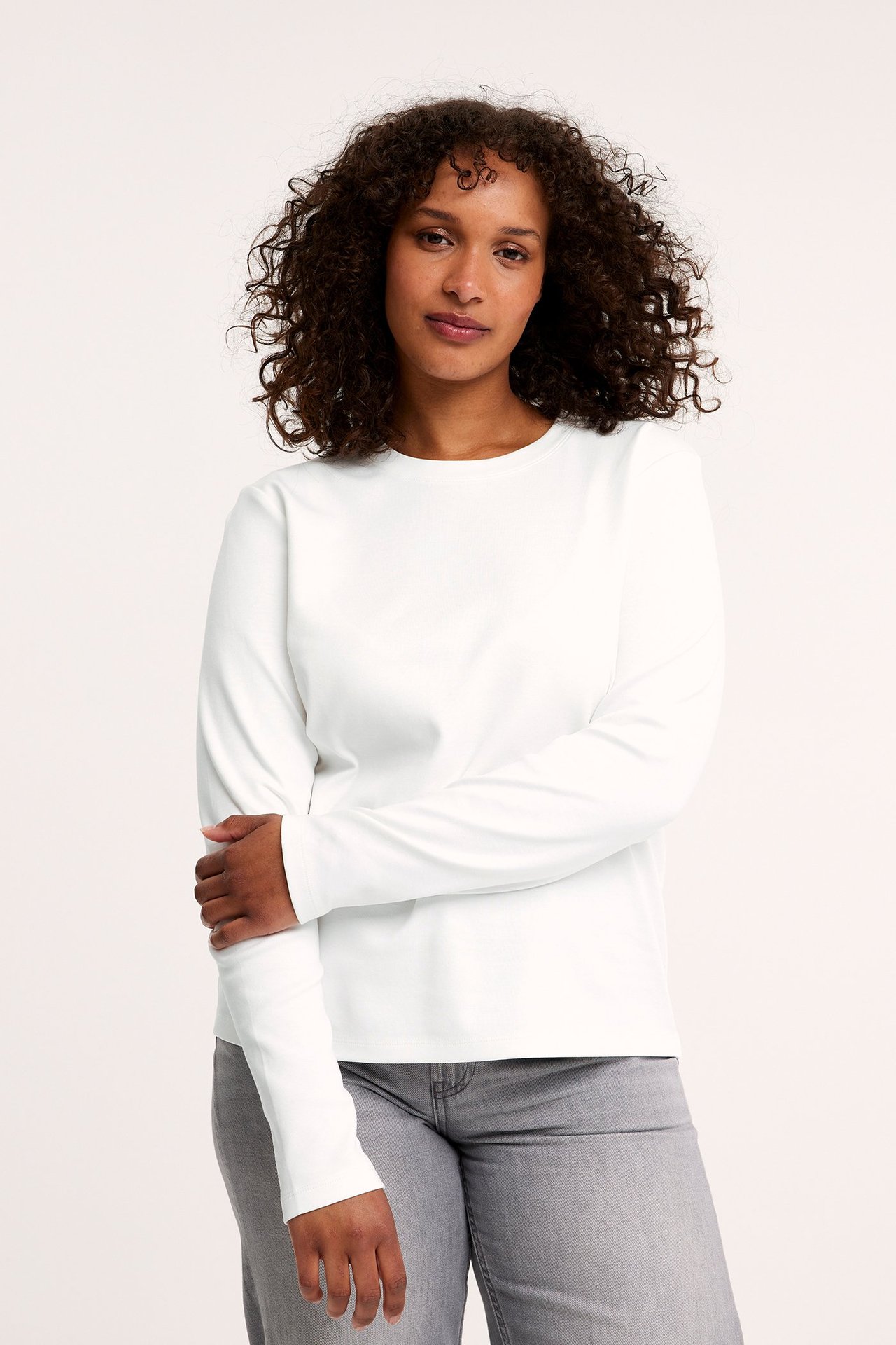 Frontbilde av en kvinne i en offwhite langermet bomulls-t-skjorte med rund hals og grå jeans.