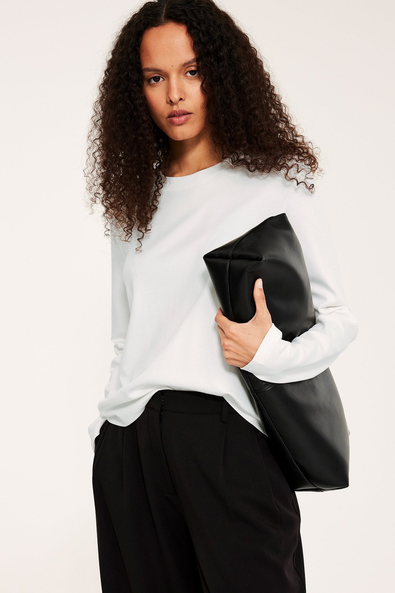 Framifrån: Kvinna i offwhite, långärmad bomullstopp, svarta byxor, håller en svart handväska.