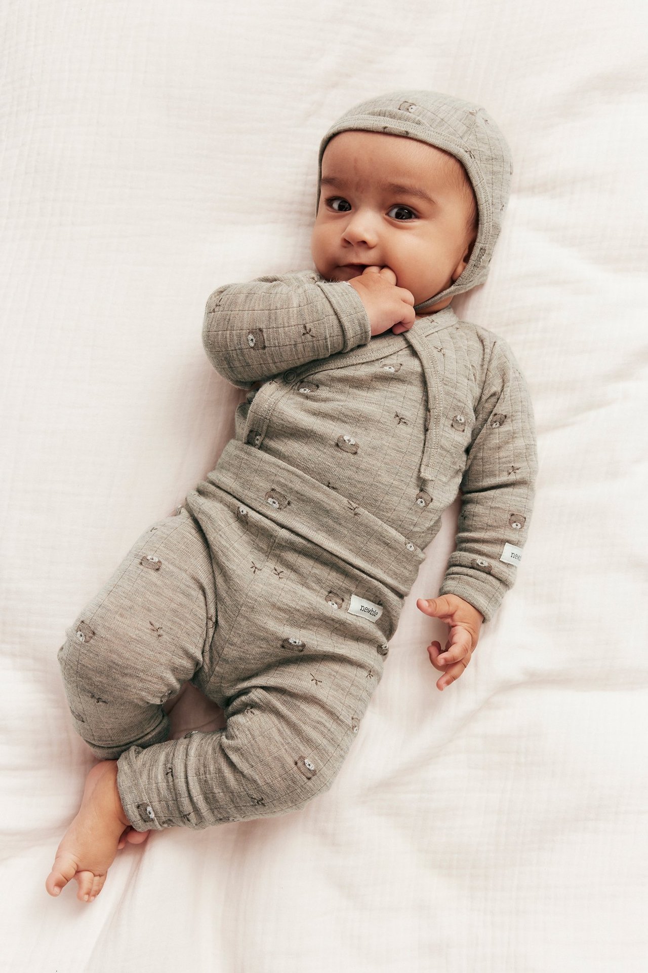 Ylhäältä kuvattuna, vauva beigenvärisissä melange-ribbineuleleggingseissä nallekuviolla, yhteensopivassa kietaisupaidassa ja myssyssä.