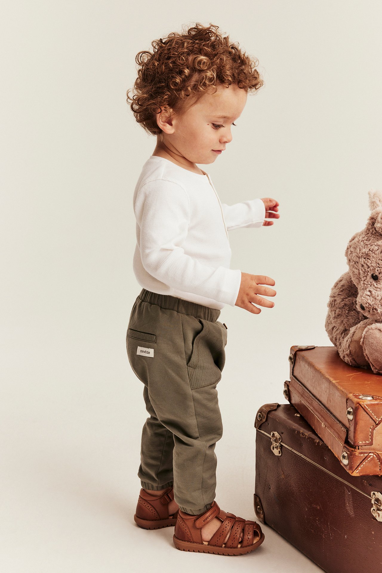 Sidovy p
R1; ett barn i vit ribbad l
R1;ng
R1;rmad body, gr
R1;na joggers och bruna sandaler.