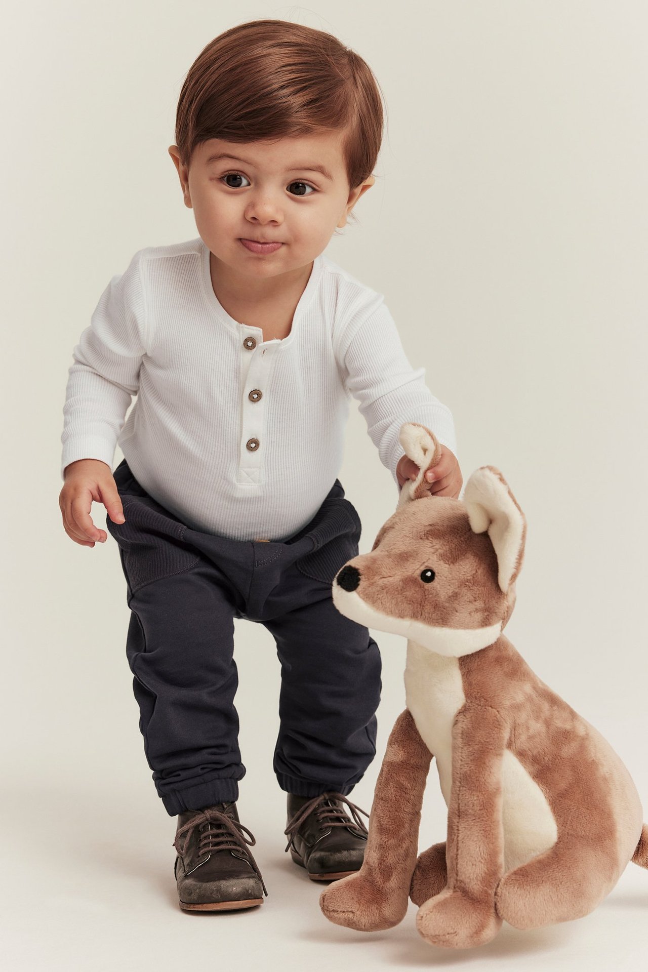 Z przodu: Niemowlę w białym prążkowanym body, ciemnych spodniach, brązowych butach, trzyma pluszowego lisa.
