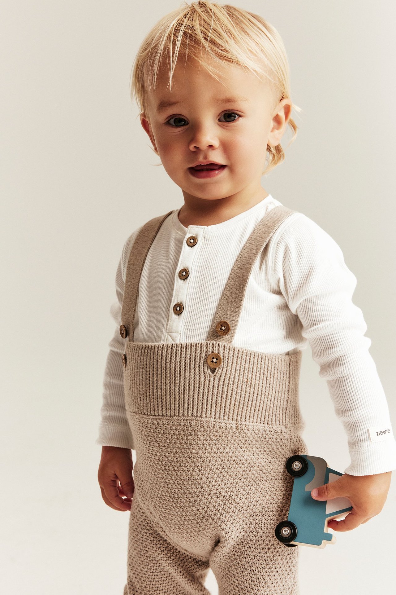 Baby i hvit ribbet langermet body med knapper, under beige strikket selebukse, holder lekebil.