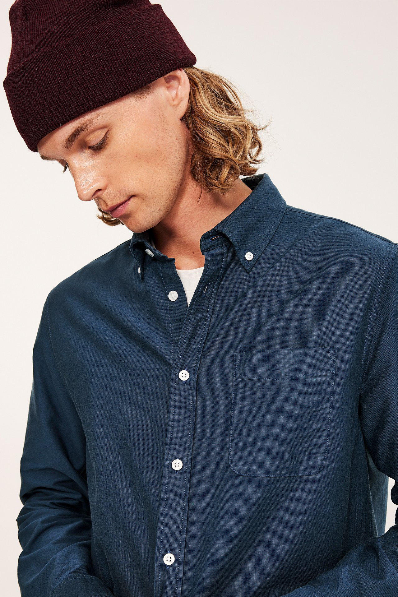 Närbild på en man i en mörkblå melerad regular fit oxfordskjorta med button down-krage och en vinröd mössa.