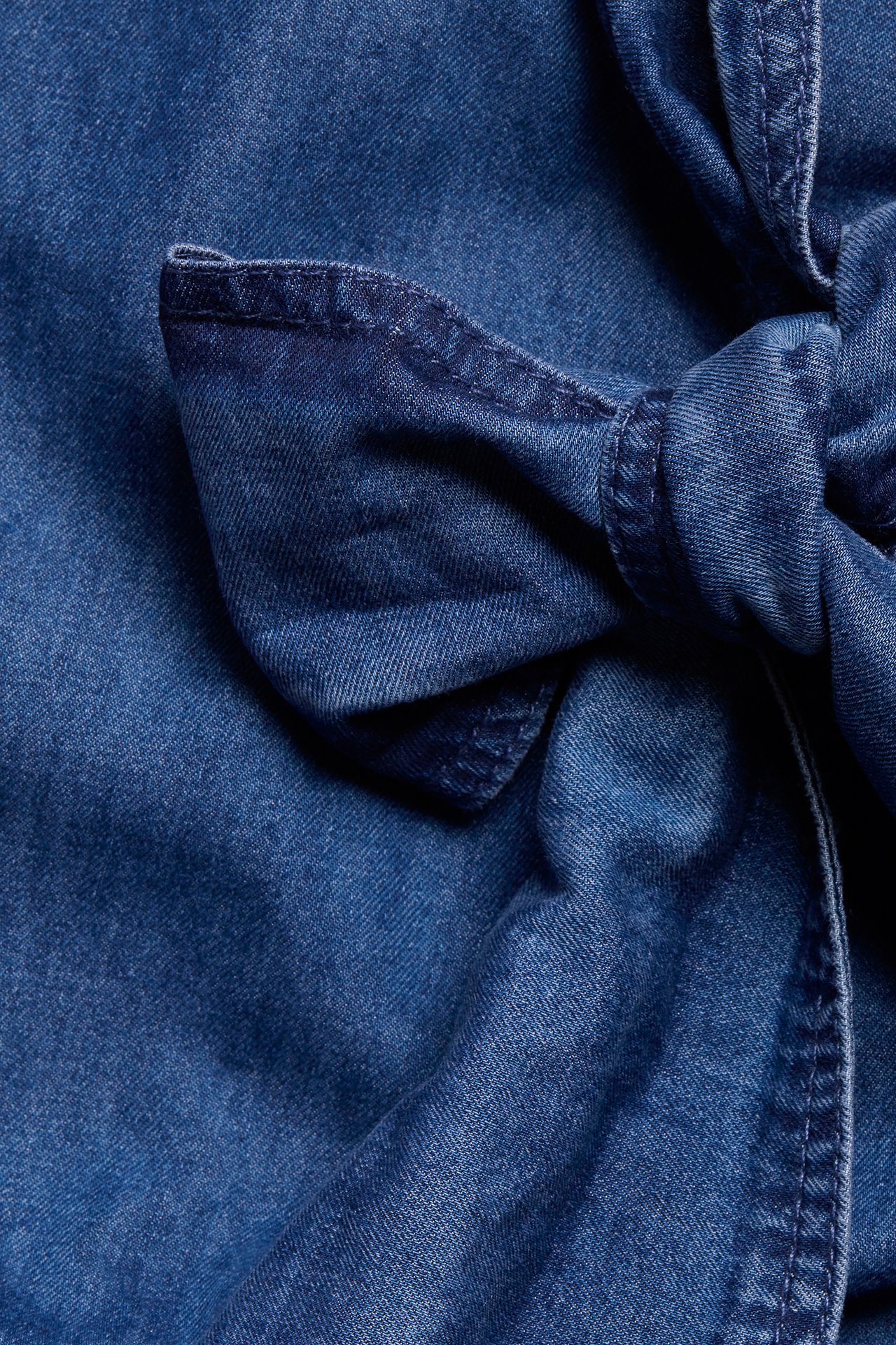 Nærbilde av mørkeblå bomulls-denimskjorte med knyttet sløyfedetalj.