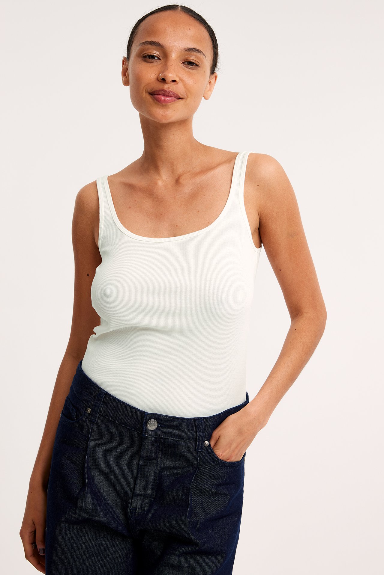 Frontvy av en kvinna i ett offwhite ribbat straplinne och mörkblå jeans.