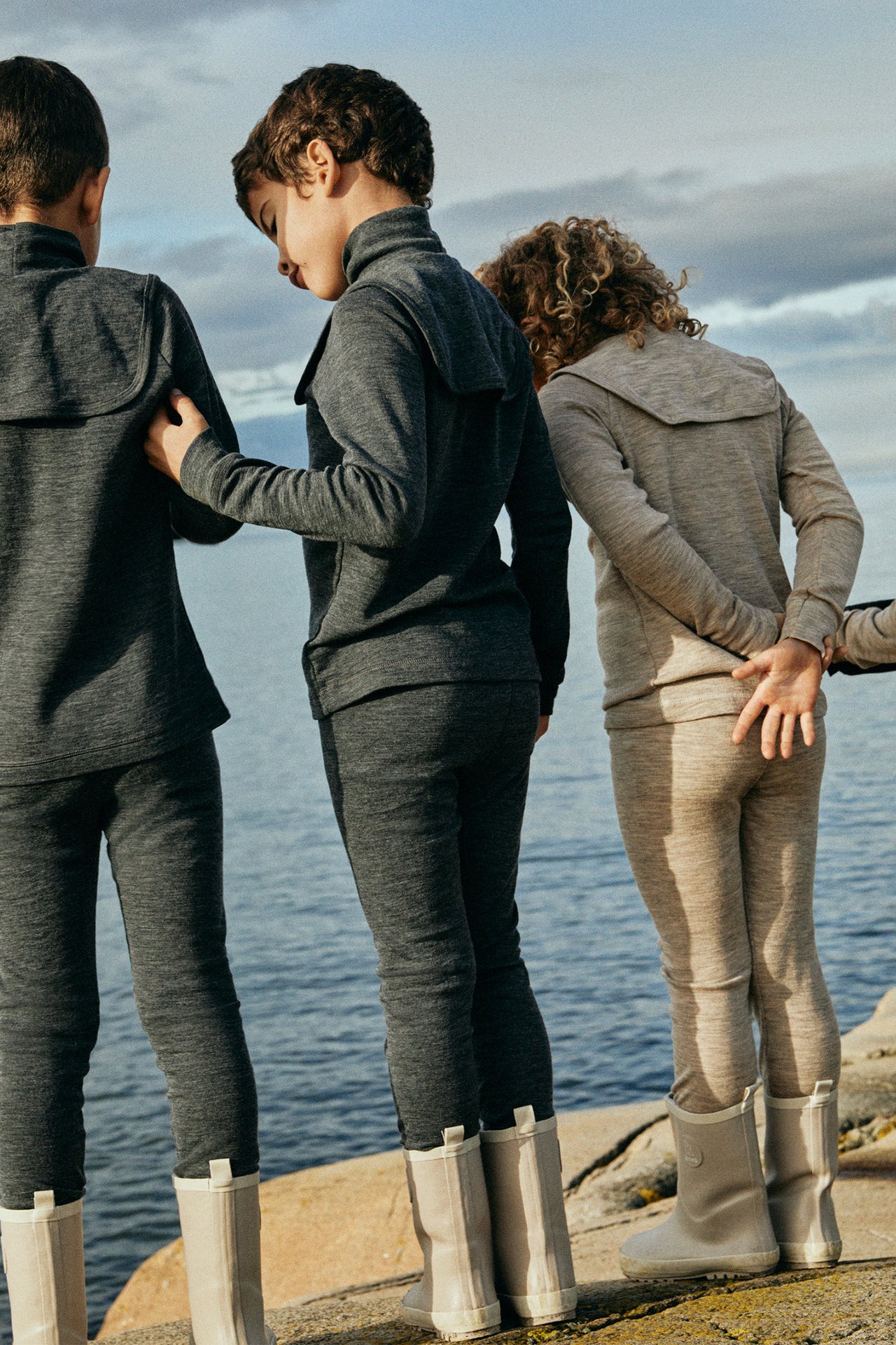 Widok z tyłu dzieci w ciemnoszarych i jasnobeżowych melanżowych kompletach bielizny z wełny merino i jasnobeżowych butach.