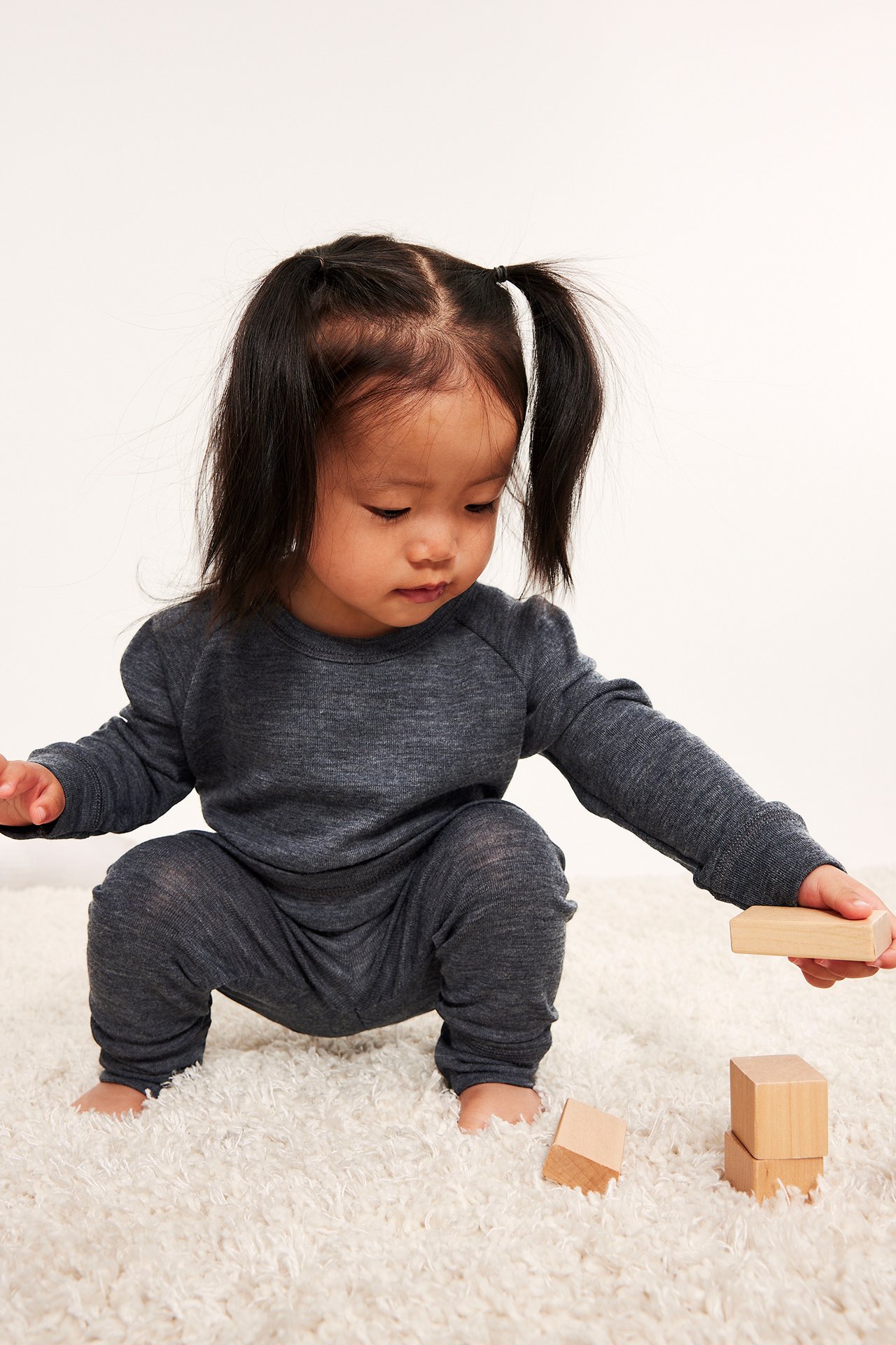 Frontvisning av en baby i mørkegråmelert merinoull underdelsbukse og matchende topp, leker med klosser.