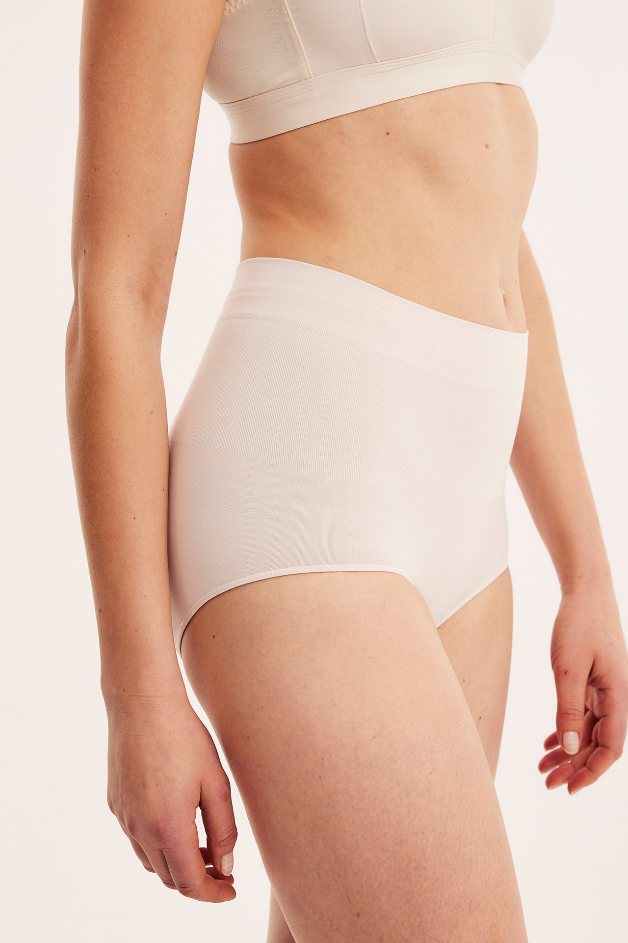 Sidebilde av en kvinne i lys beige sømløs shapingtruse med høy midje og matchende BH.