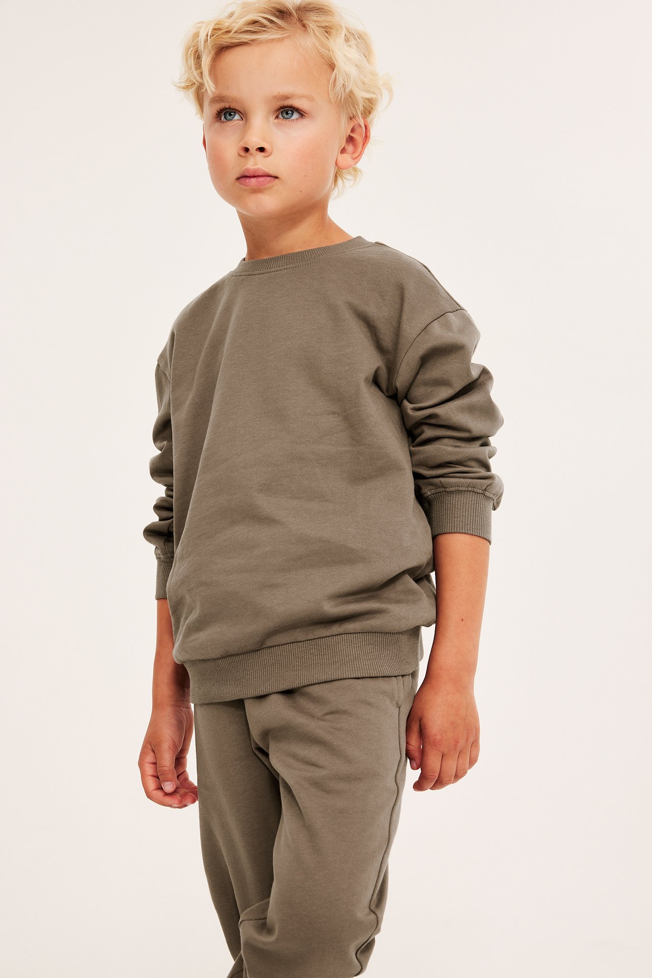 Framifrån, ett barn i en lös, enfärgad brun bomullssweatshirt och matchande mjukisbyxor.