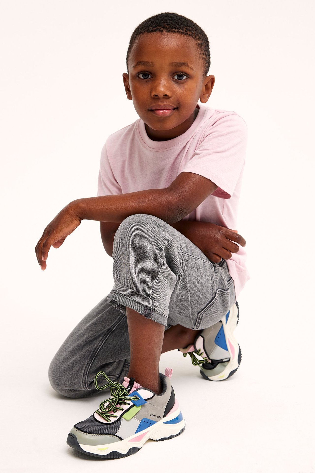 Widok z przodu klęczącego dziecka w jasnoróżowym bawełnianym T-shircie, szarych podwiniętych dżinsach i wielokolorowych sneakersach.