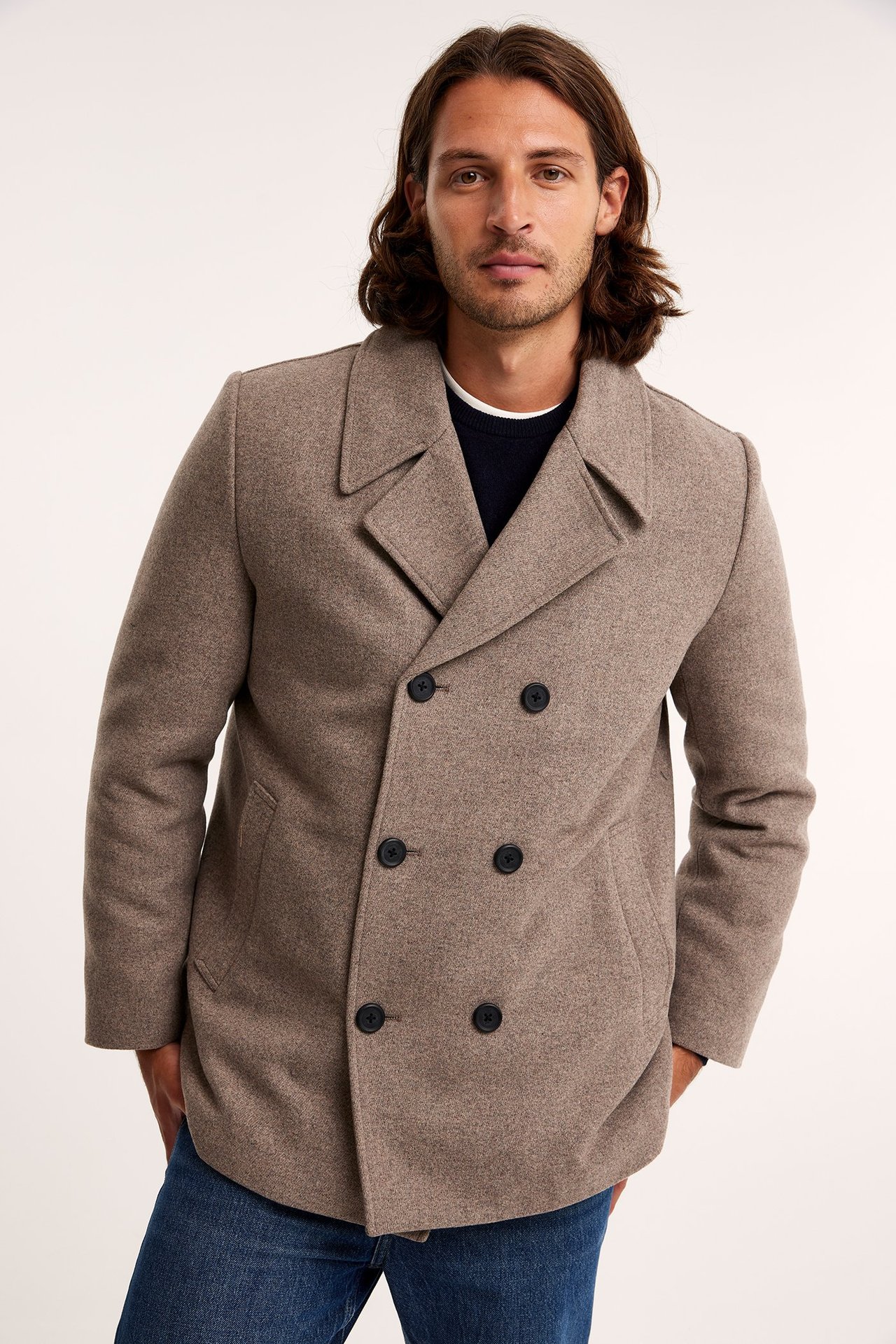 Widok z przodu: Mężczyzna w beżowej melanżowej dwurzędowej kurtce, ciemnoniebieskim swetrze i dżinsach.